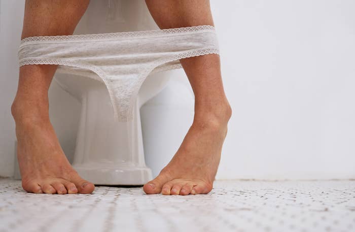 Person sitting on a toilet with underwear pulled down to their ankles, feet shown on a tiled bathroom floor