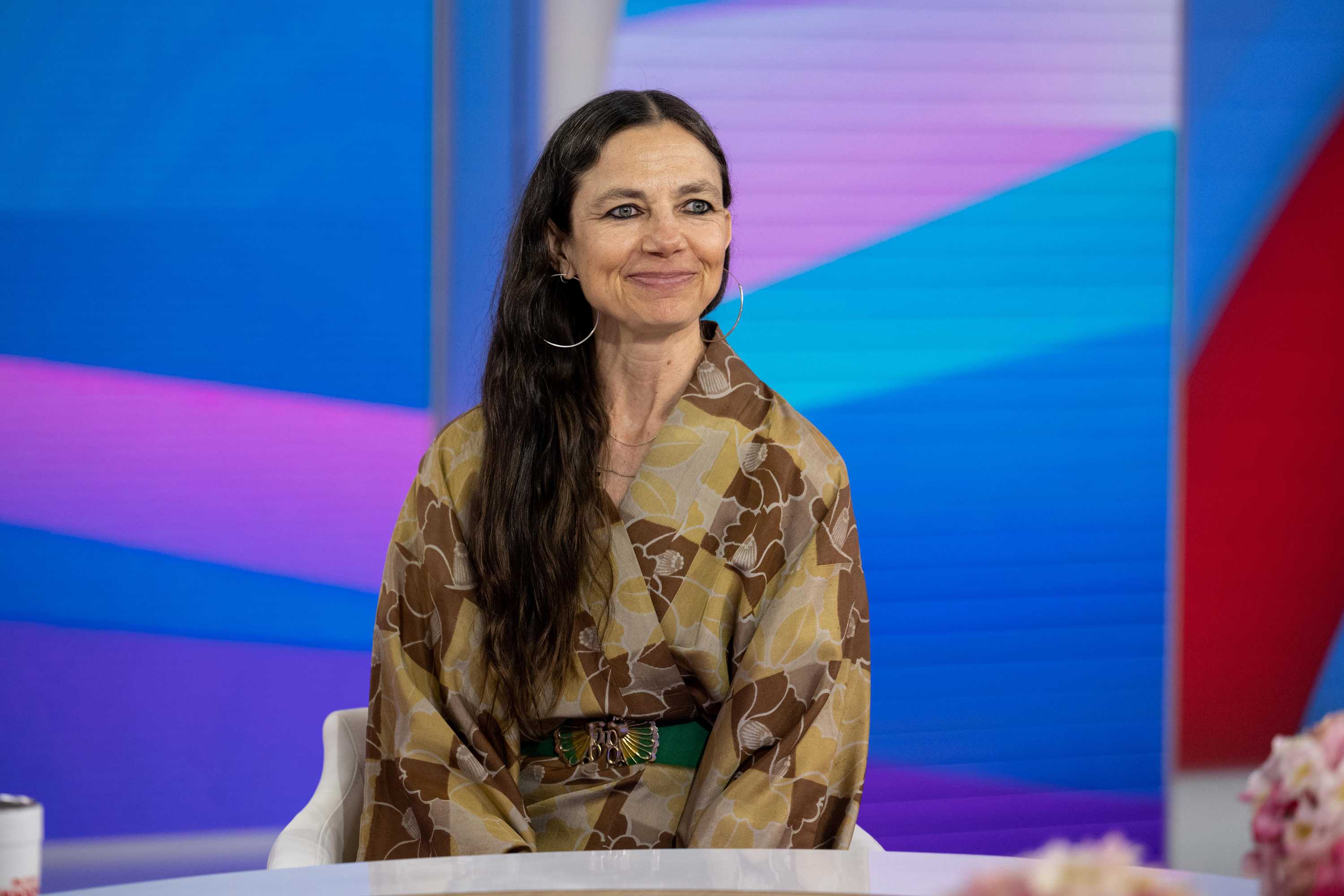 Justine Bateman in a patterned blouse sits smiling on a talk show set featuring a colorful, abstract background