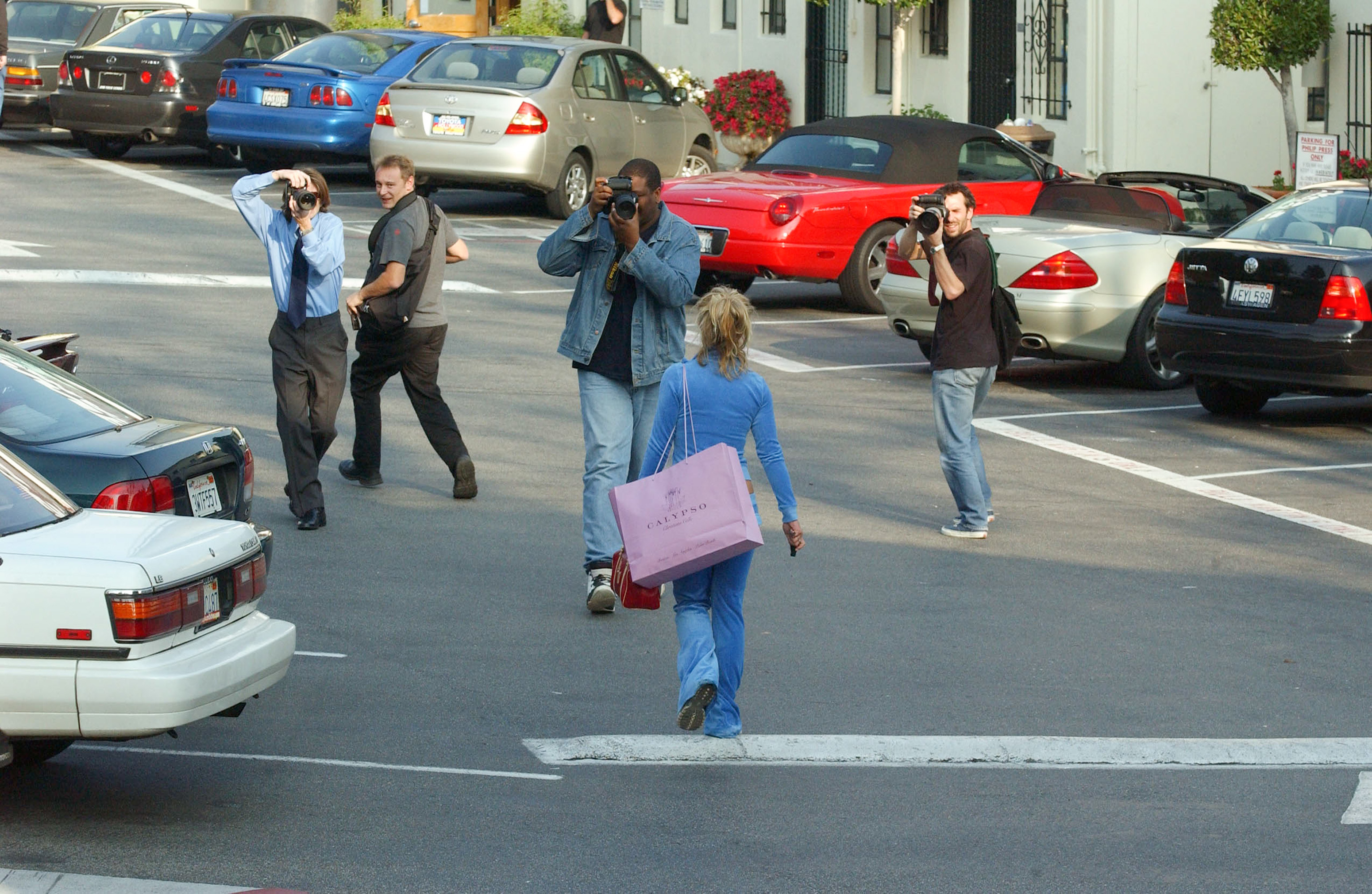 Person walking in a parking lot being photographed by paparazzi. The person has a pink shopping bag and is wearing a casual outfit