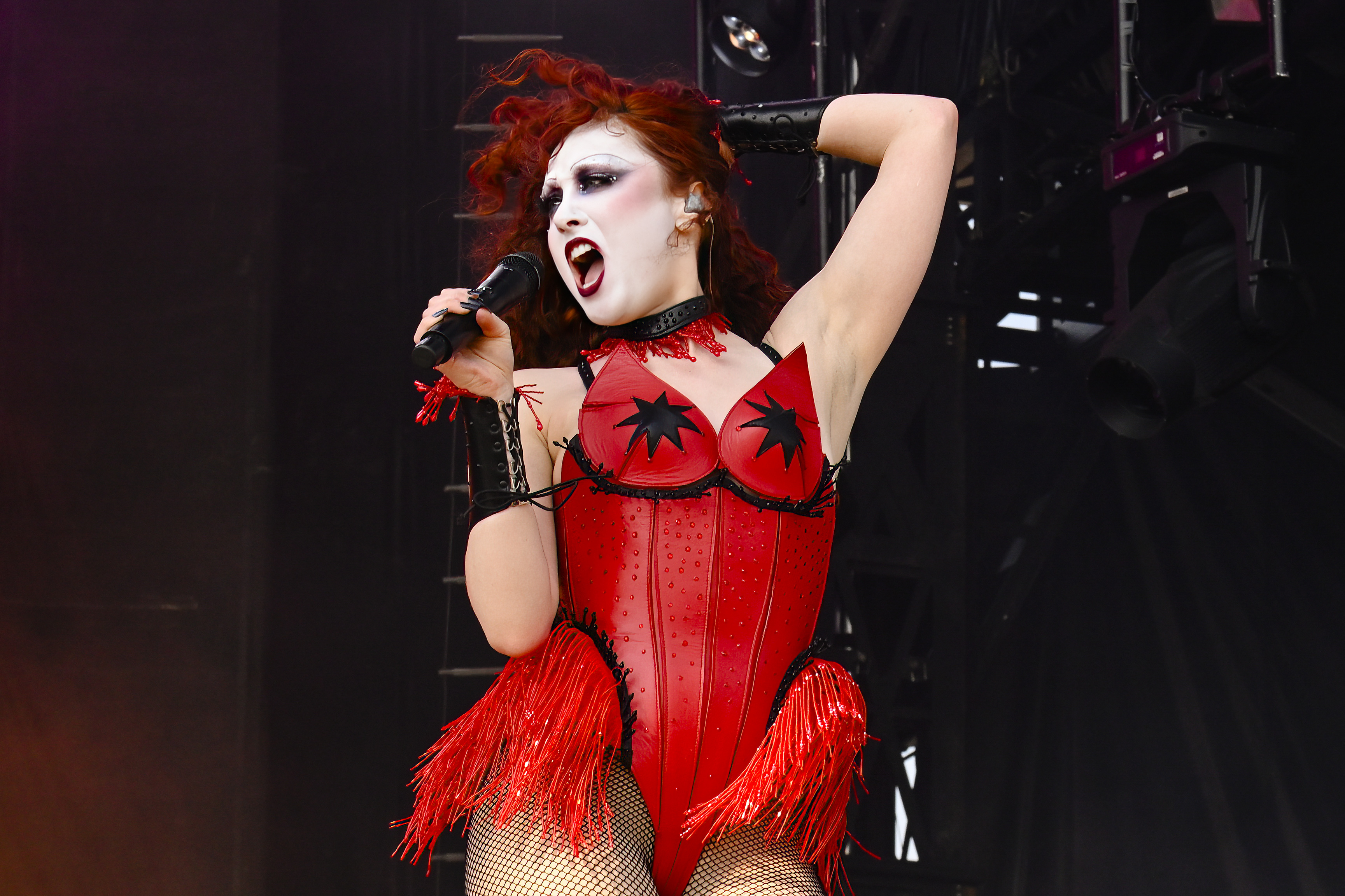Chappell Roan performing on stage, wearing a corset with star-shaped details, fringed accents, and fishnet stockings, holding a microphone