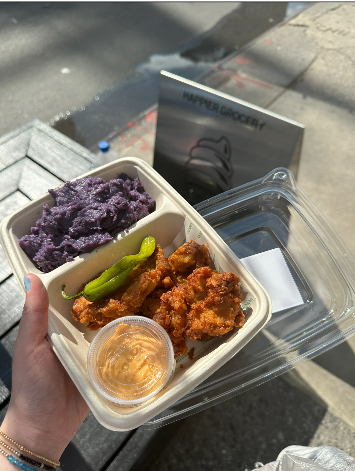A hand holding a takeout container with fried chicken, a green pepper, purple mashed potatoes, and a small cup of dipping sauce, outside of a grocery store
