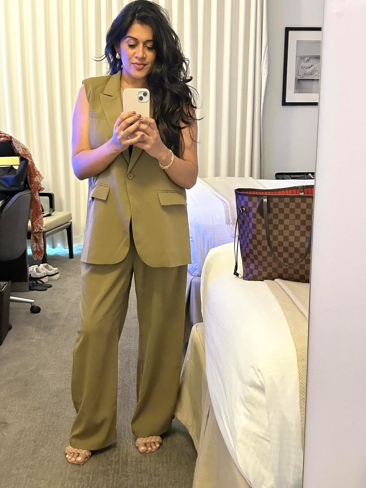 A reviewer takes a mirror selfie in a hotel room, dressed in a loose-fitting, sleeveless suit and holding a smartphone. A designer handbag is seen on the bed