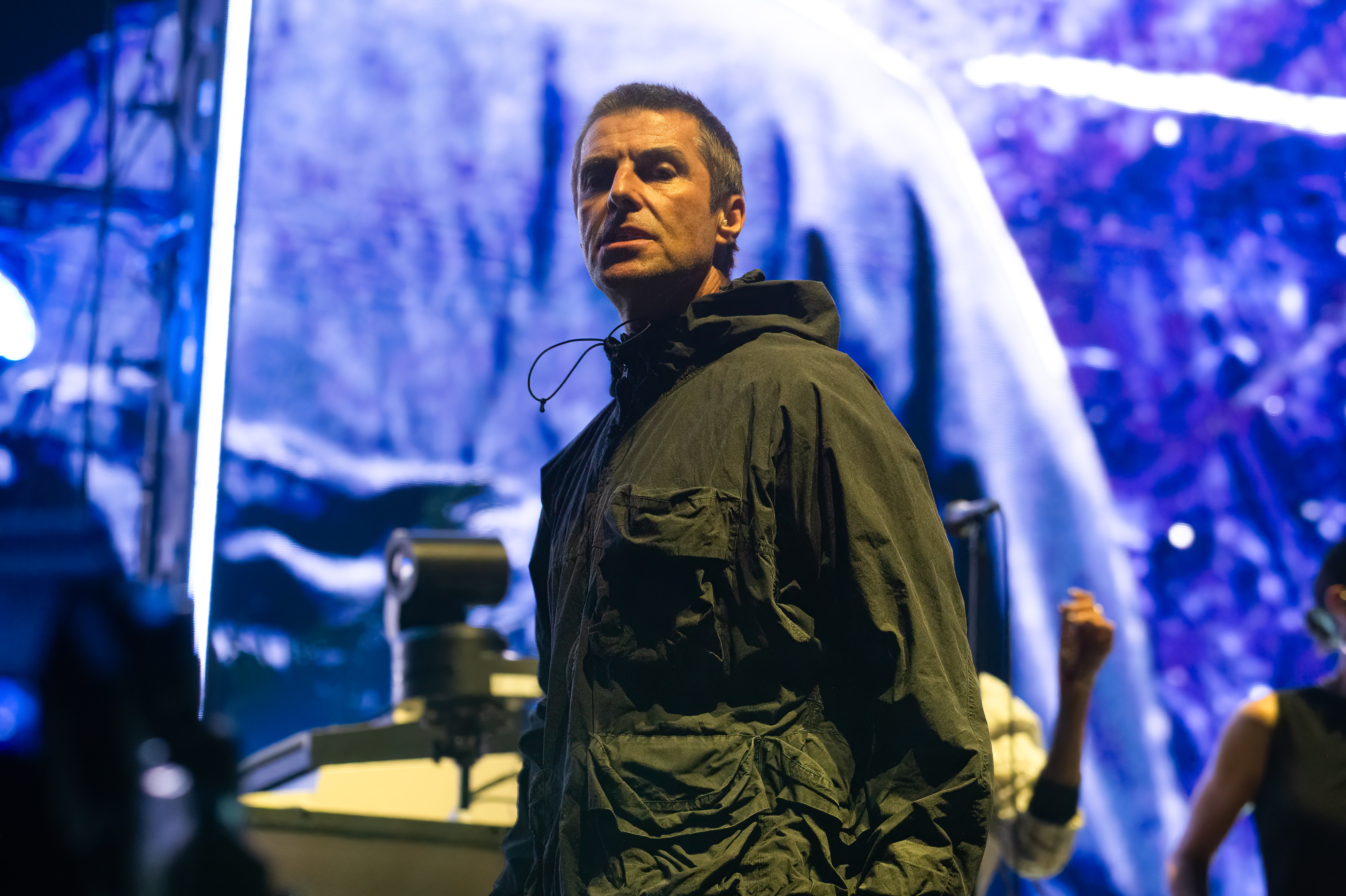 Liam Gallagher performs on stage, wearing a casual jacket. Bright, abstract backdrop behind him