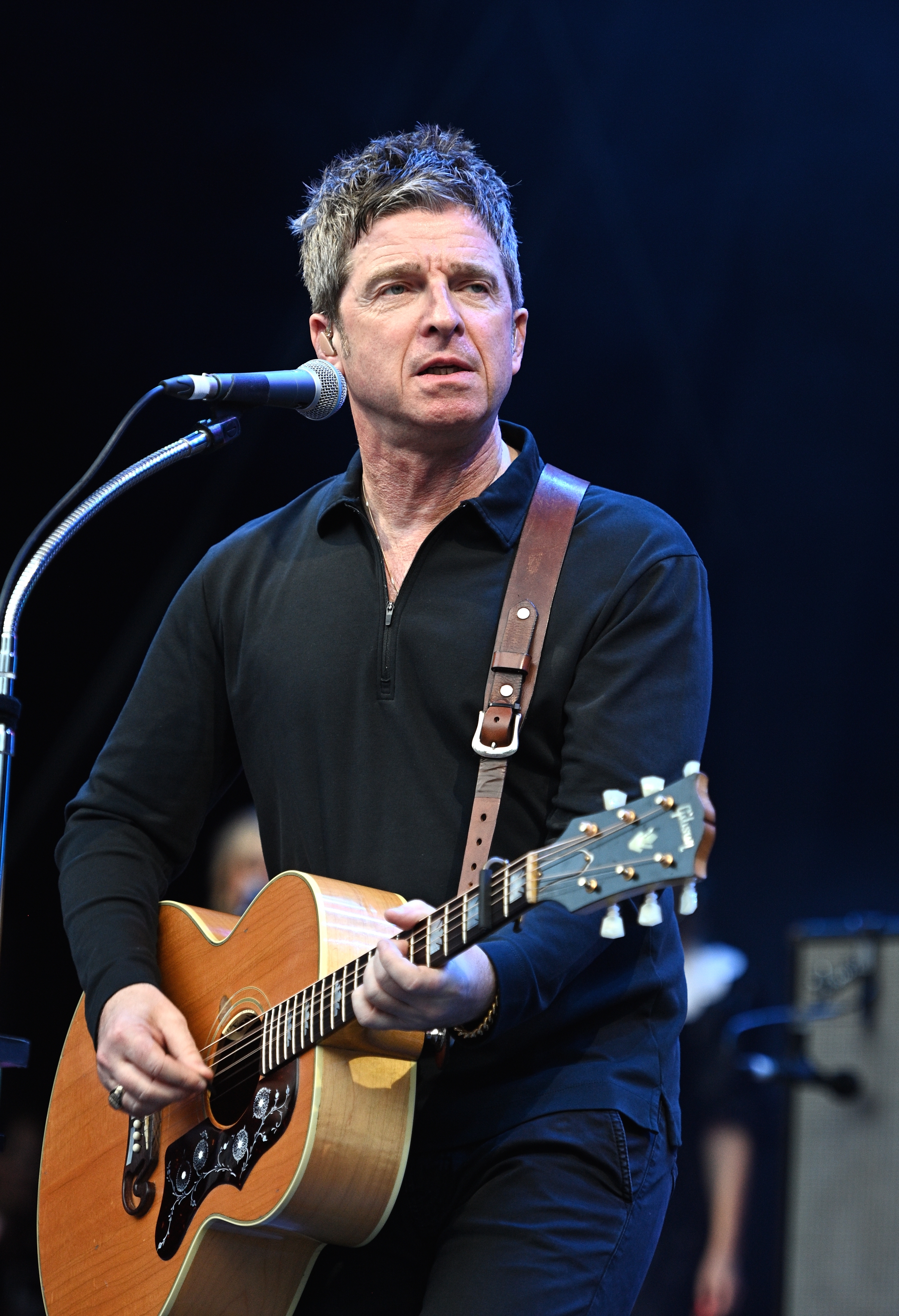 Noel Gallagher playing an acoustic guitar on stage, wearing a casual black shirt