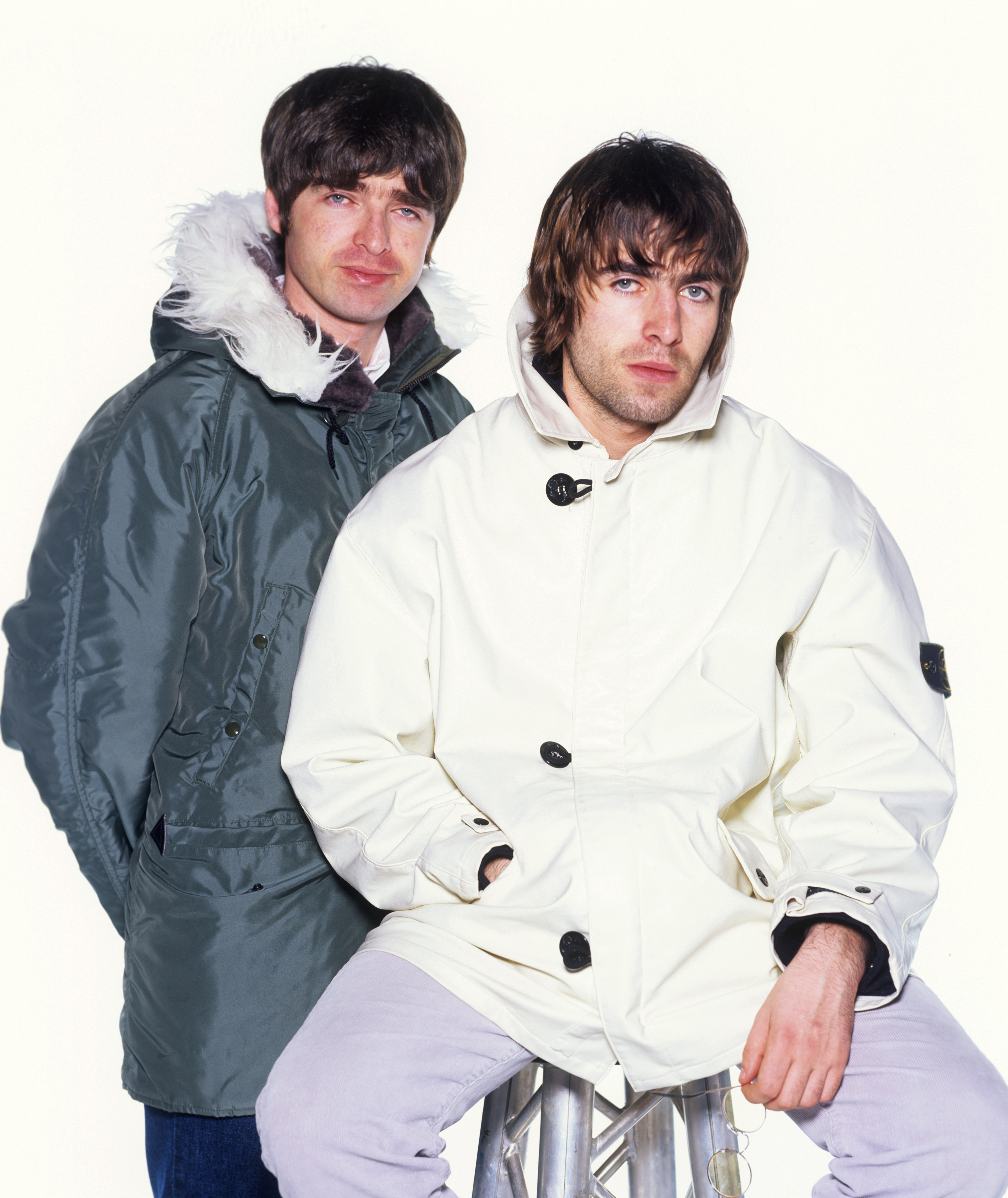 Noel Gallagher and Liam Gallagher, both wearing large jackets, pose together with neutral expressions