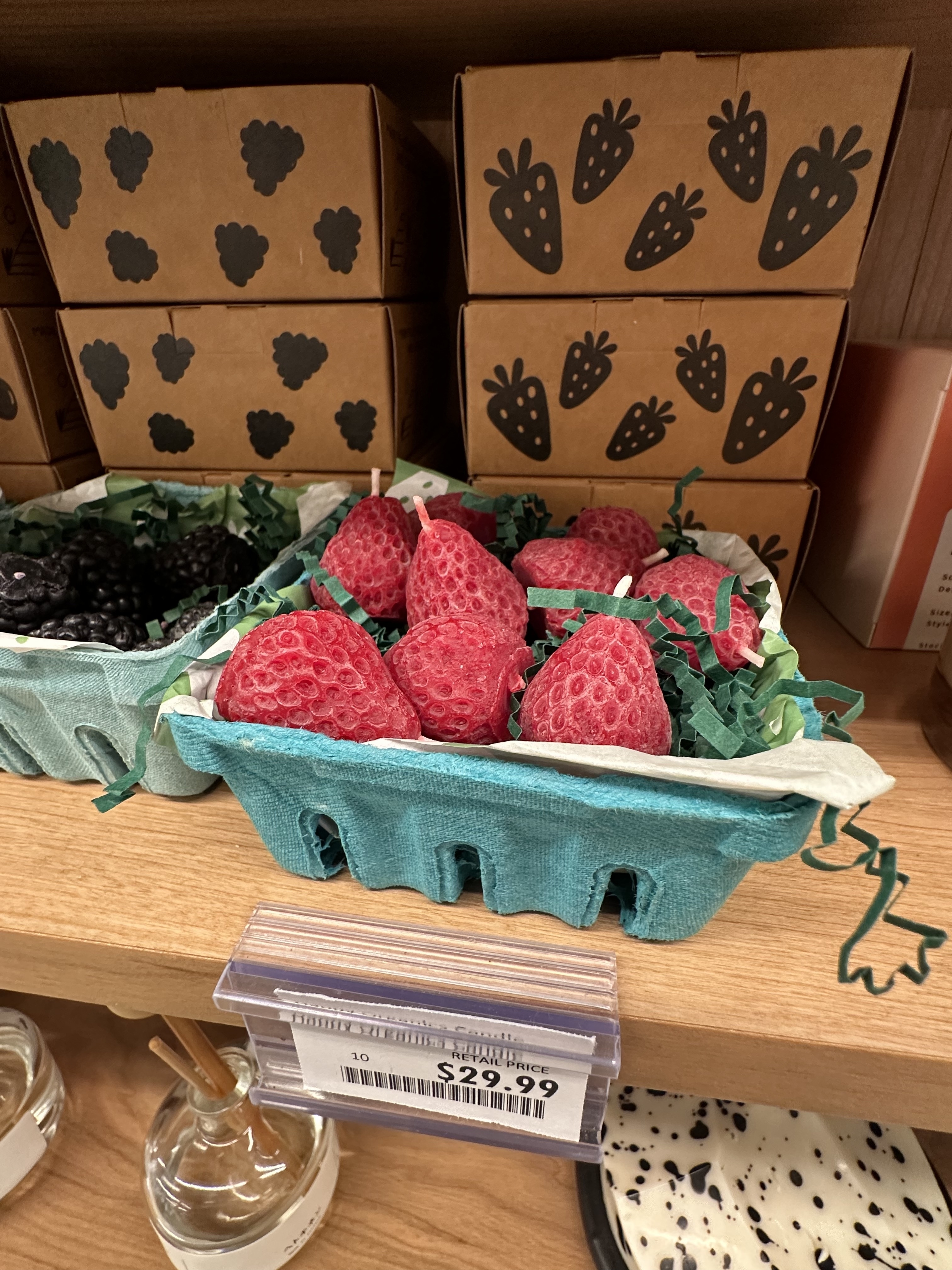 Pint container of realistic soap strawberries priced at $29.99 on a store shelf, with cardboard boxes featuring strawberry designs in the background