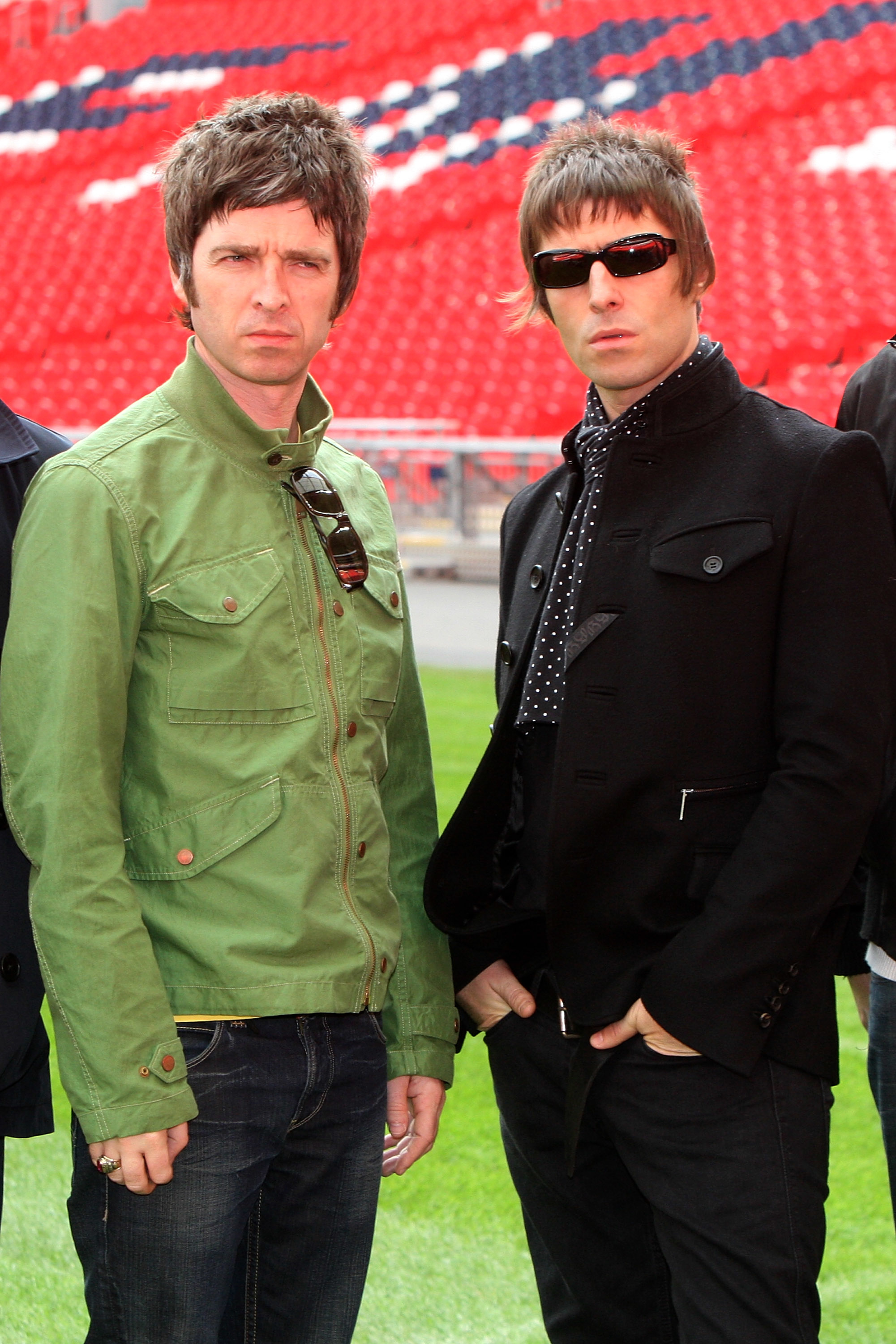 Noel Gallagher and Liam Gallagher standing in front of stadium seats. Noel wears a casual jacket, and Liam sports sunglasses and a stylish jacket