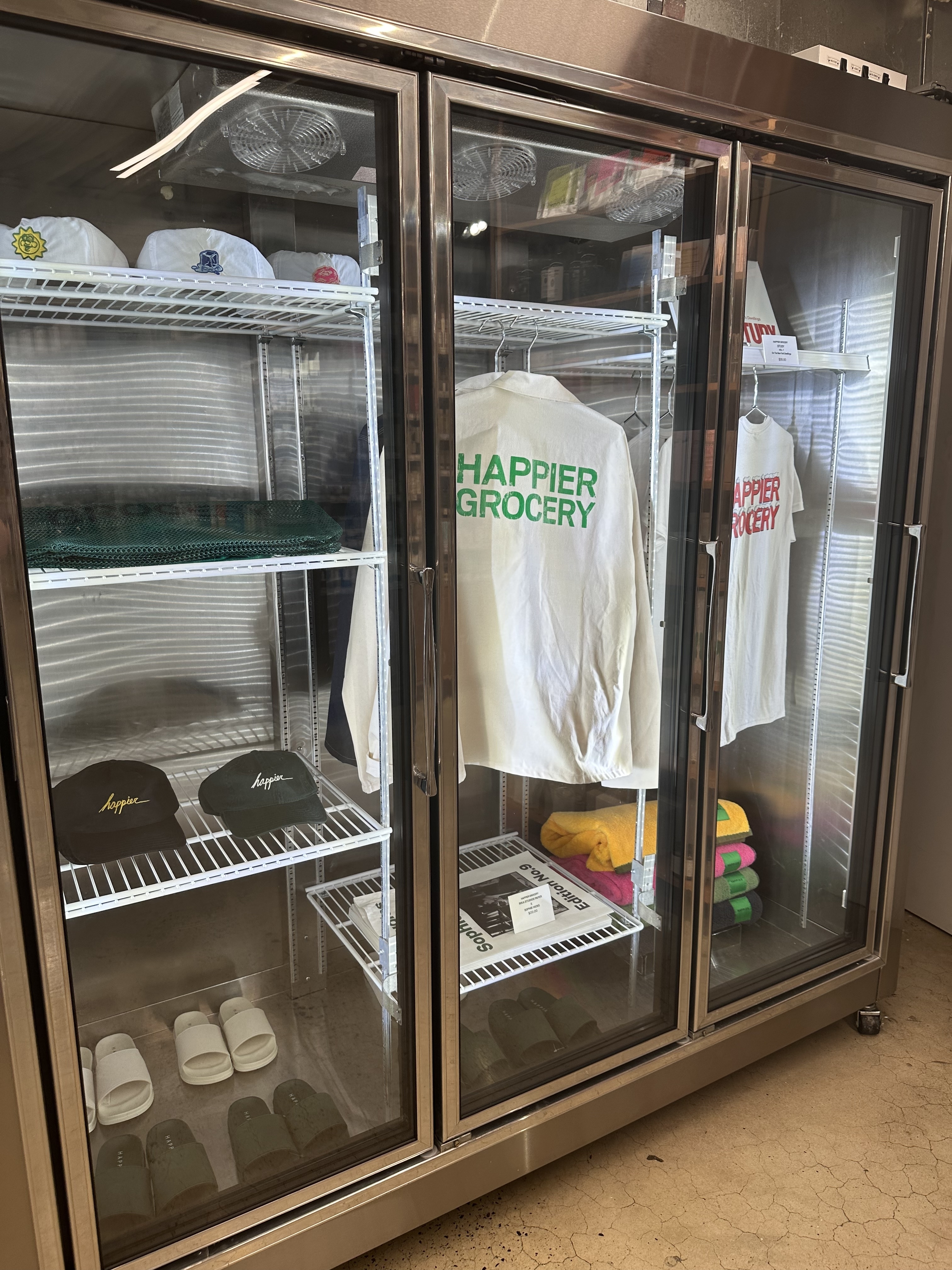 Refrigerated display case with hats, shirts, slippers, and various clothing items branded with "Happier Grocery."