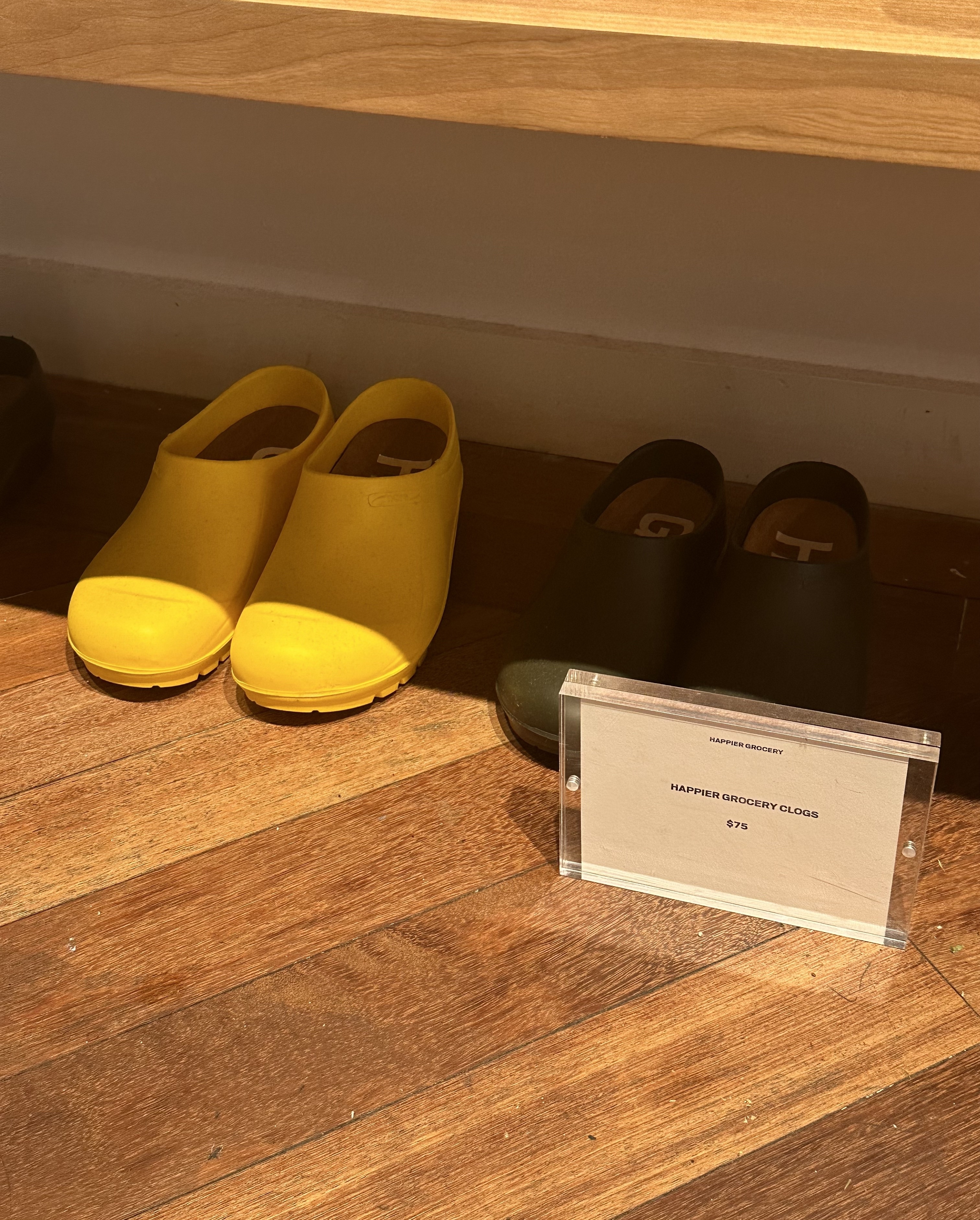 Two pairs of rubber clogs, one yellow and one dark green, are displayed on a wooden floor. A sign reads, "NAPPIES GROGGY CLOGS GT, SIZE 5."