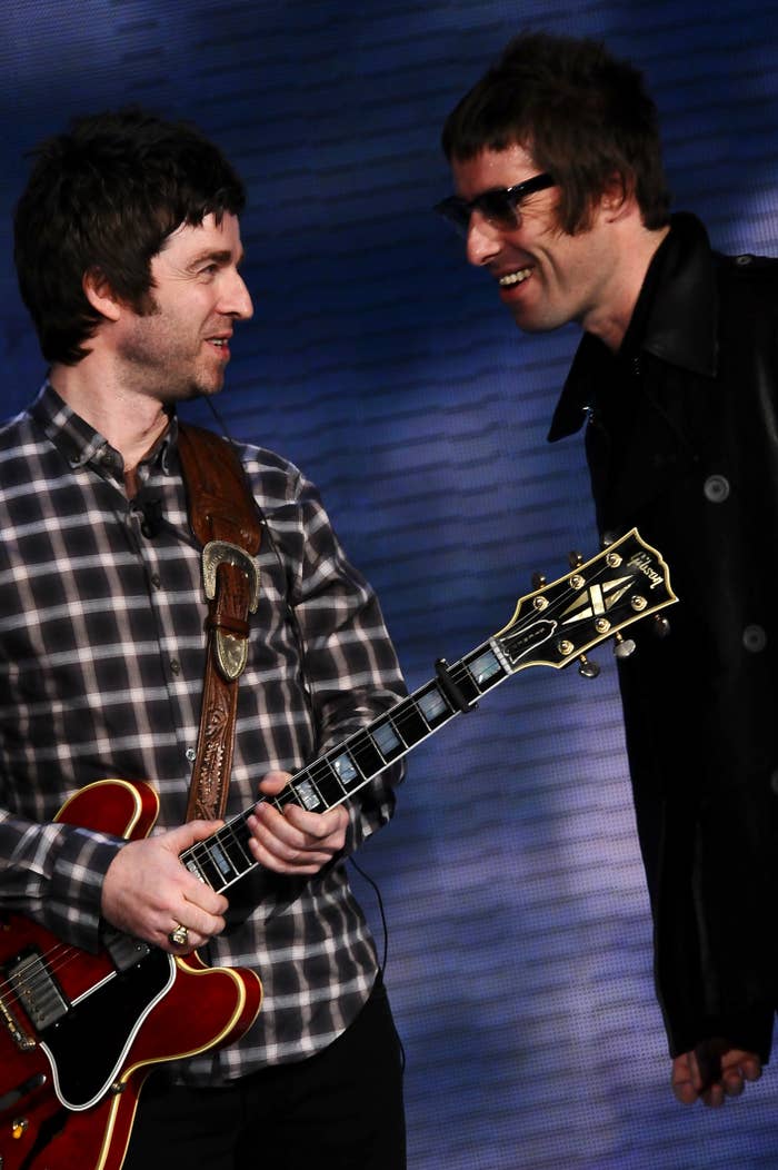 Noel Gallagher and Liam Gallagher standing and smiling at each other; Noel holding a guitar, Liam wearing sunglasses and a leather jacket
