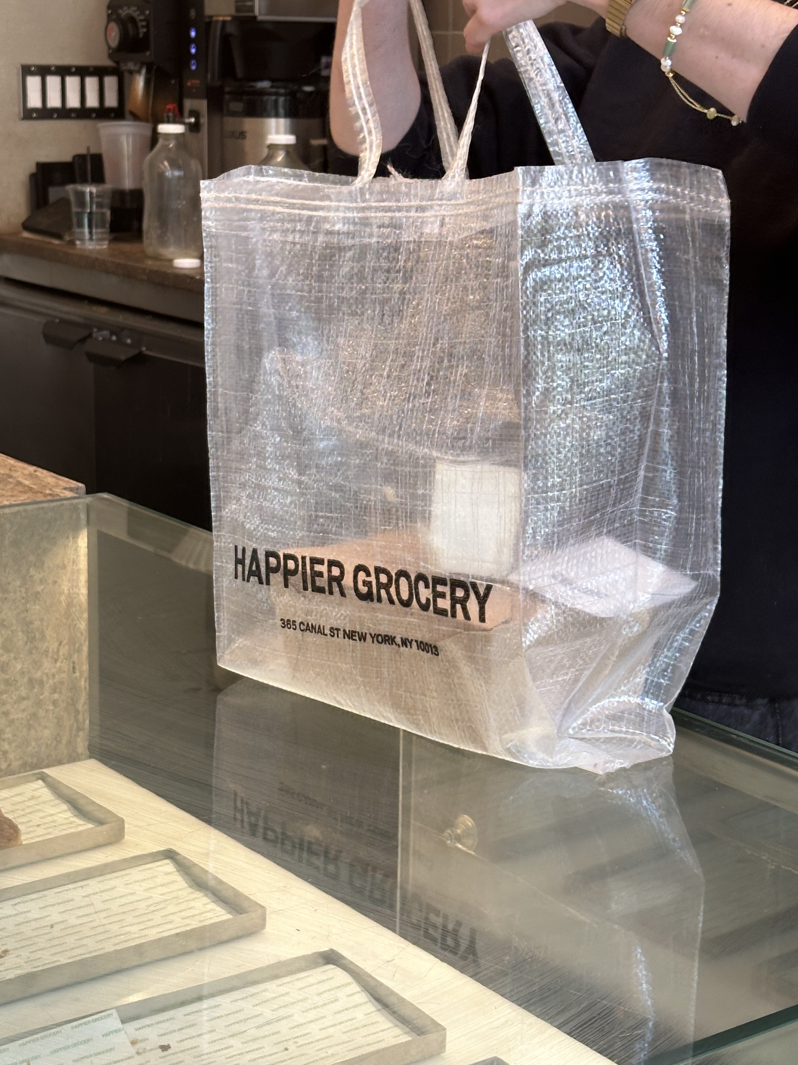 A person carries a reusable grocery bag with the text "Happier Grocery, 365 Canal St, New York, NY 10013" at a countertop inside a store