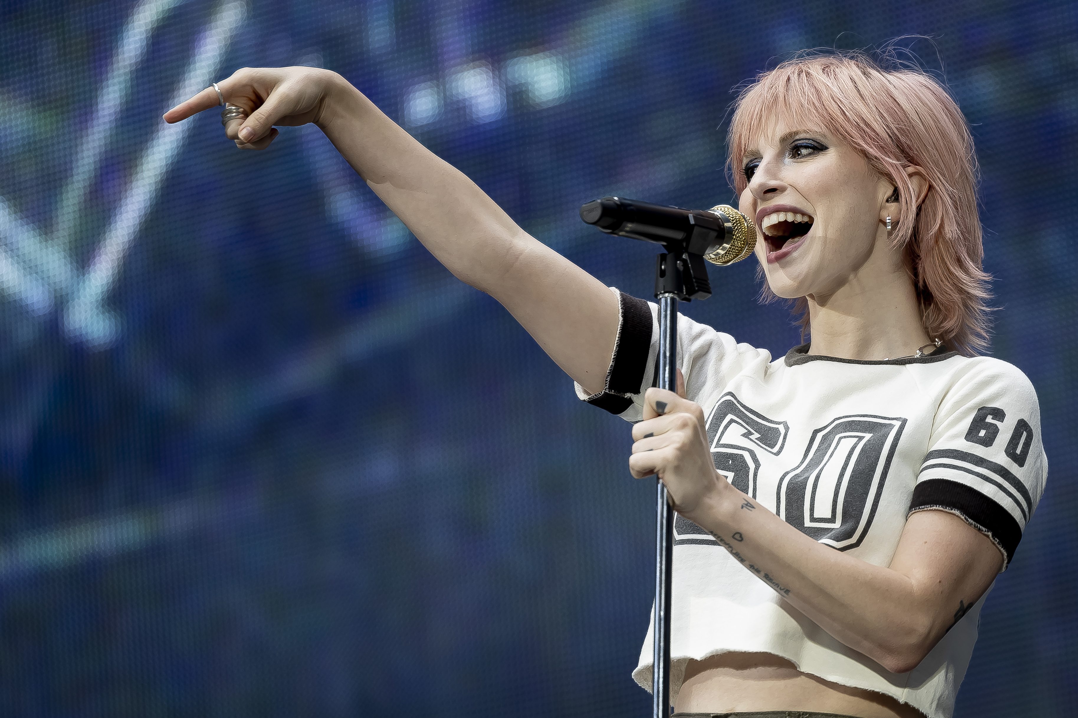 Hayley Williams performing on stage, wearing a cropped sports jersey with the number 60. She is pointing and smiling with a microphone in hand