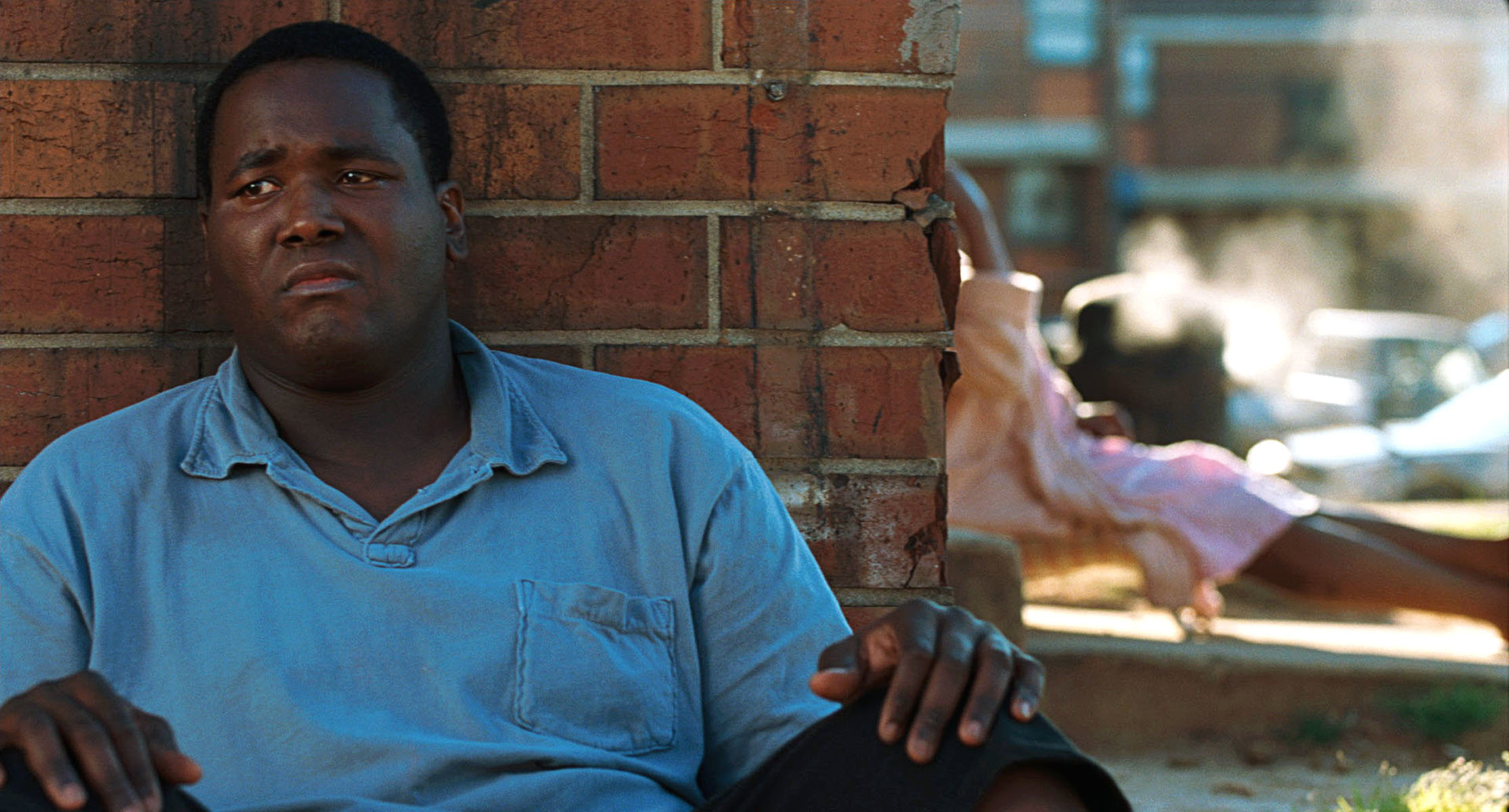 A man sits against a brick wall with a sad expression while another person lays beside a wall in the background