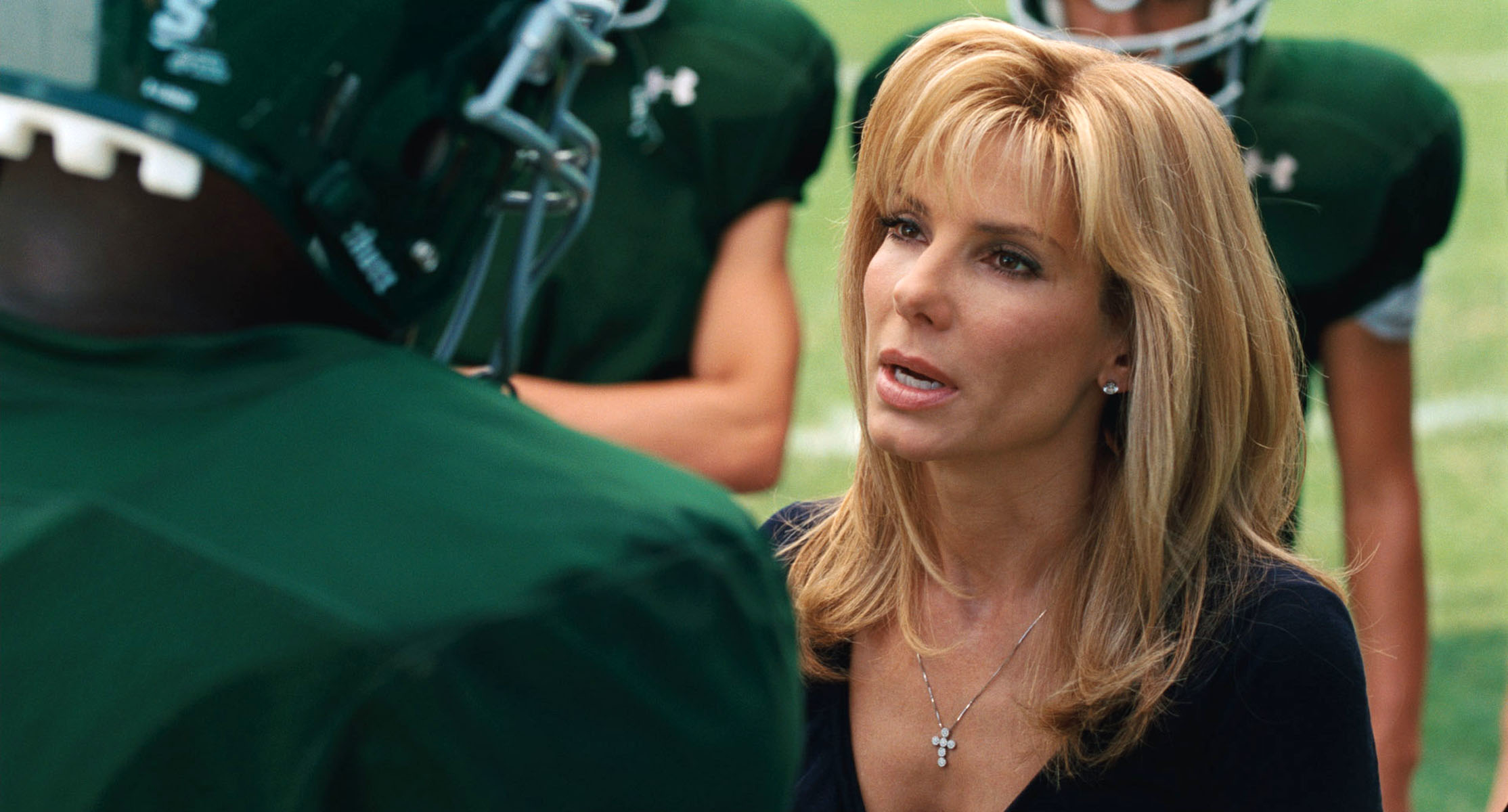 Sandra Bullock, dressed casually, talks to a football player wearing his helmet on a grassy field. Two other players stand in the background