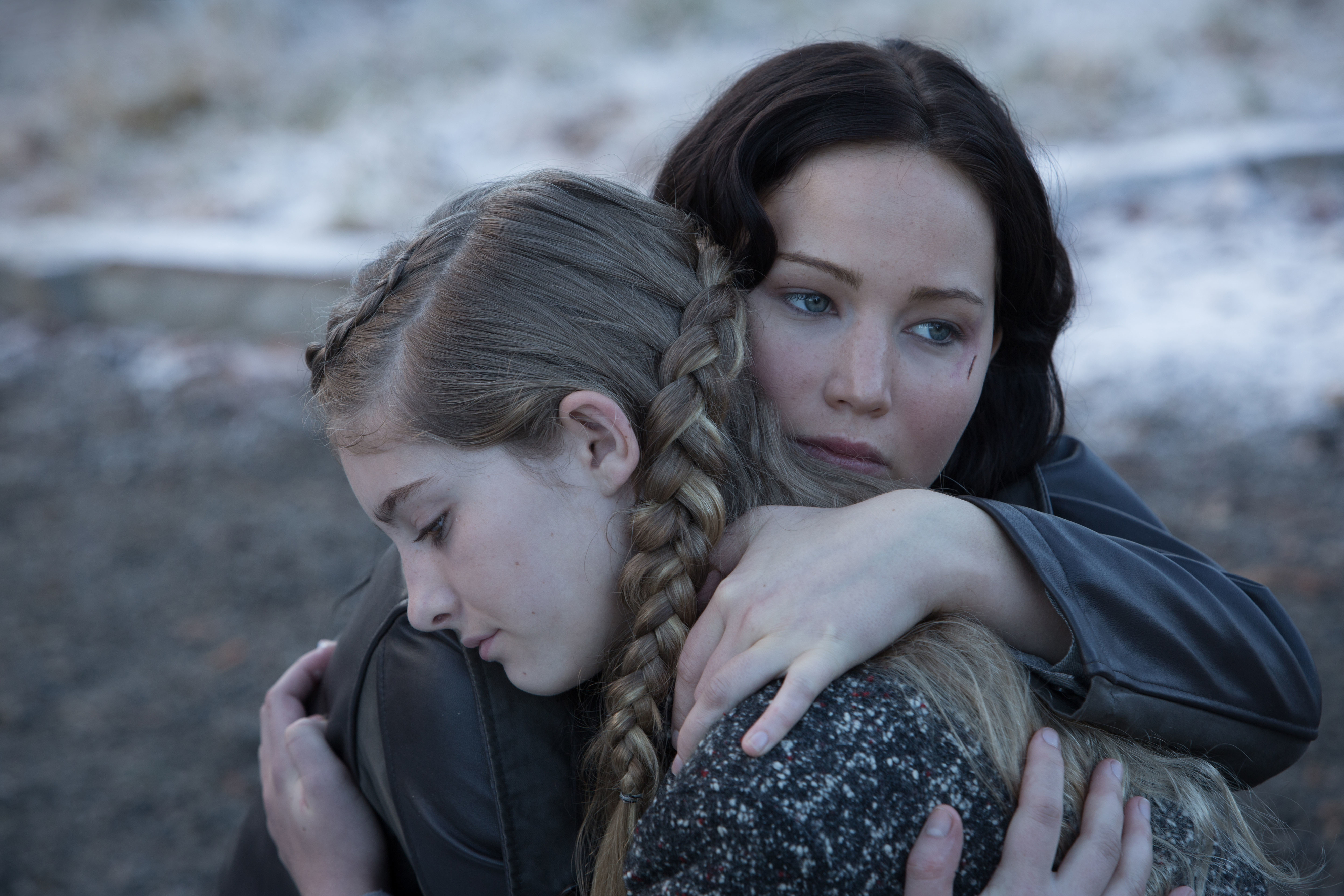 Jennifer Lawrence hugs Willow Shields while both look serious in an outdoor setting; both wear plain, practical attire likely suited for rough conditions
