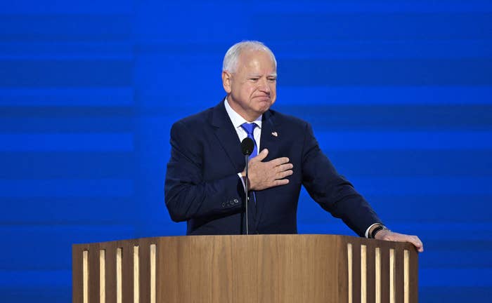 Tim Walz stands behind a podium, holding his hand to his chest while giving a speech