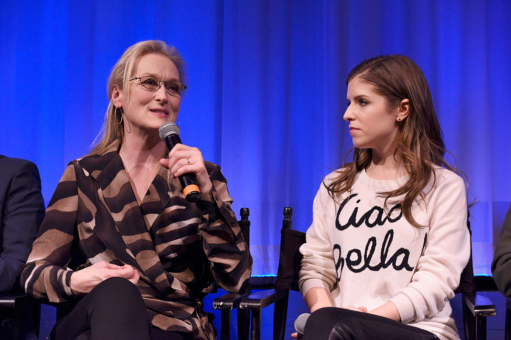 Meryl Streep in a patterned blouse speaks into a microphone while Anna Kendrick in a casual "Ciao Bella" sweater listens beside her