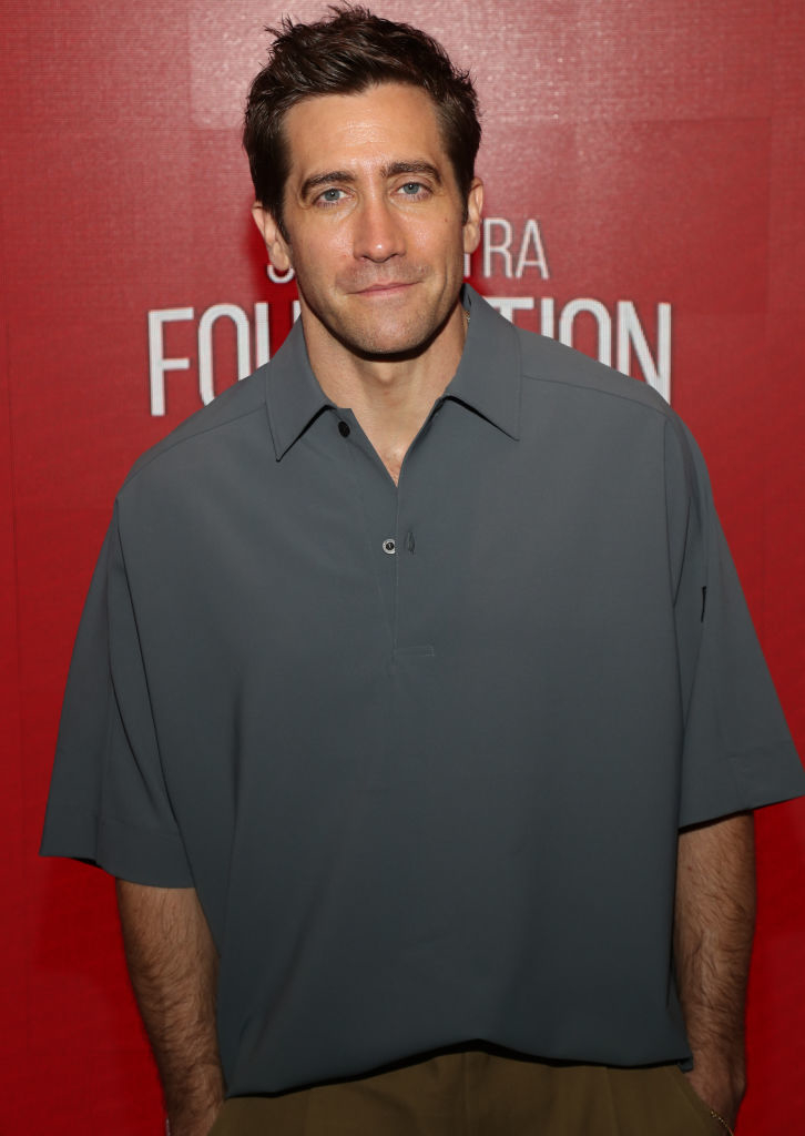 Jake Gyllenhaal on a red carpet, wearing a loose-fitting gray shirt and brown pants, smiling at the camera