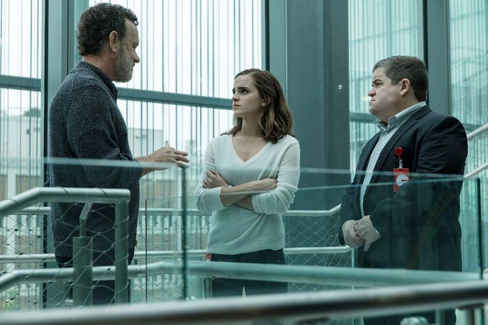 Tom Hanks, Emma Watson, and Patton Oswalt in a serious conversation on a glass-railed staircase set, all wearing casual attire