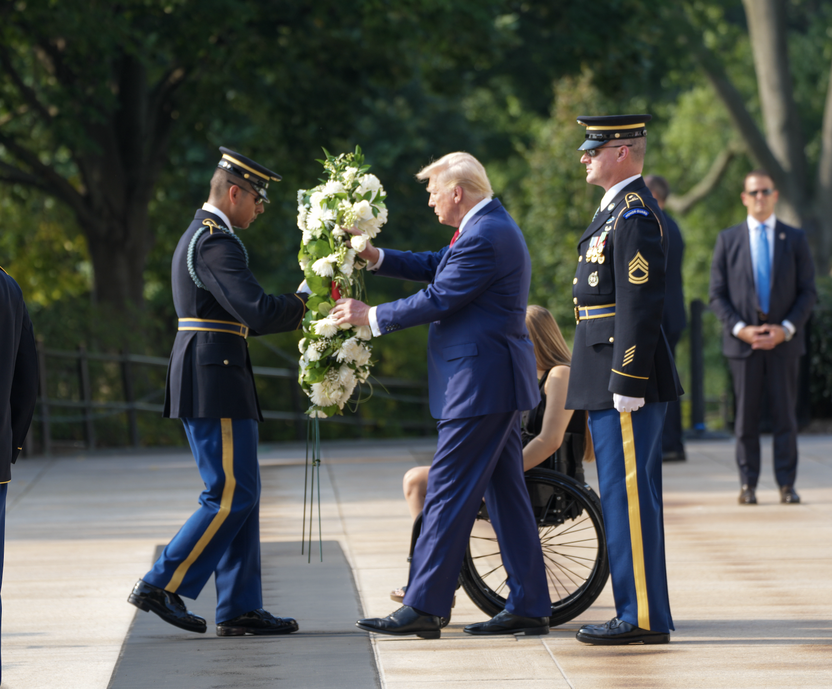 Donald Trump, in a suit, lays a wreath with two uniformed soldiers. A woman in a wheelchair and a man in a suit are in the background