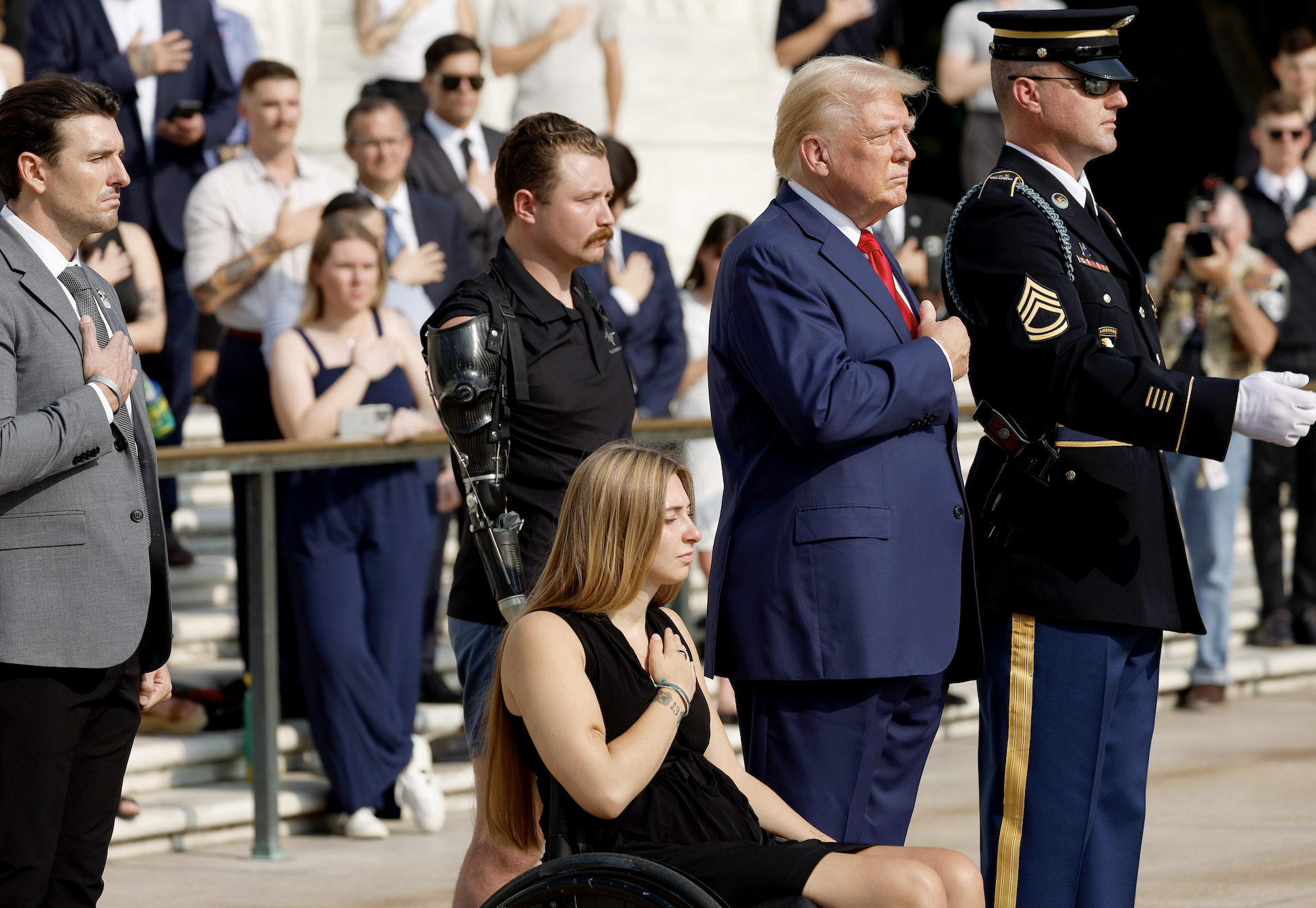 Donald Trump standing with a group, including a woman in a wheelchair and a man with a prosthetic arm, during a ceremony with hands over their hearts