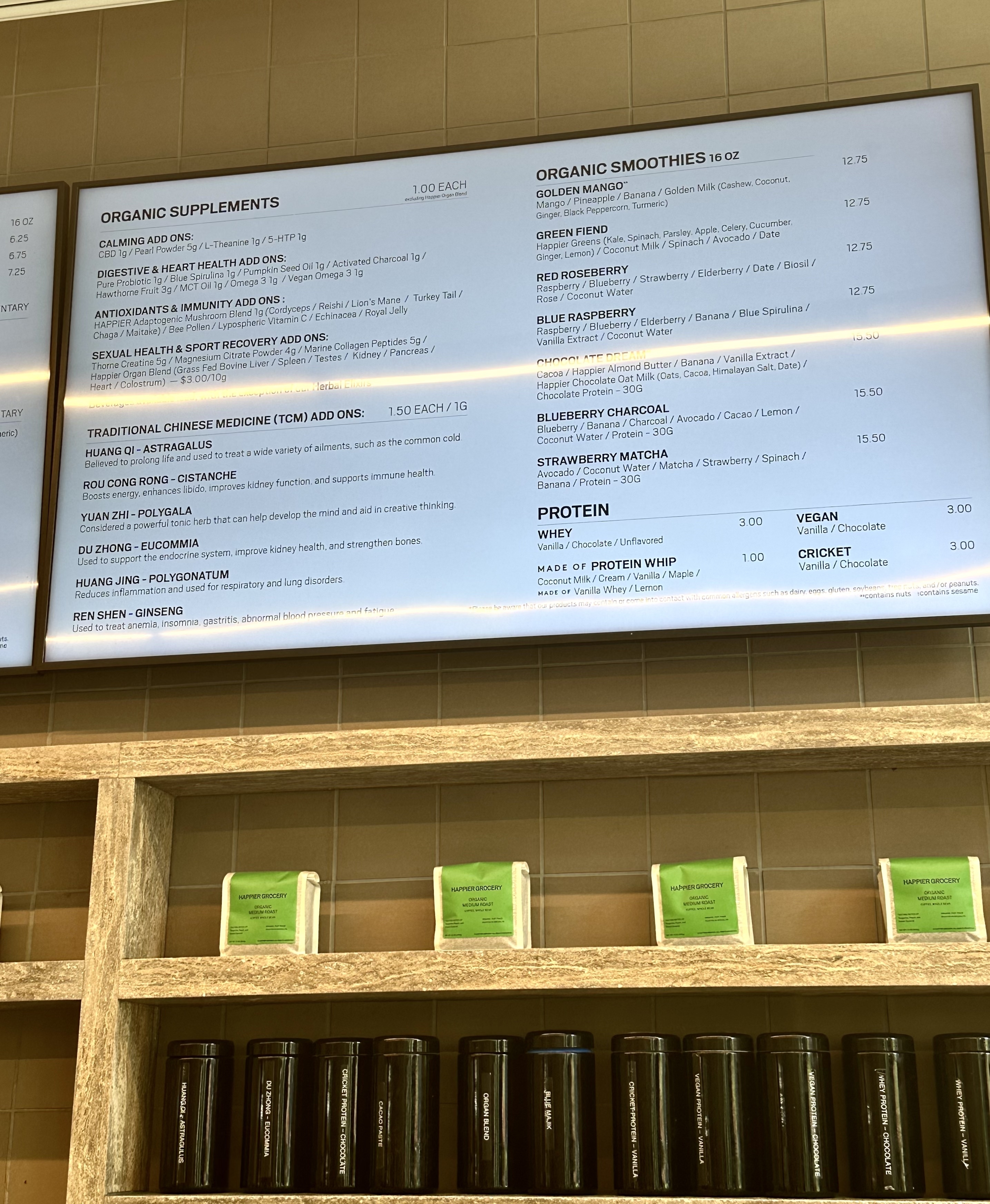 Menu board at a health food shop, showing various organic smoothies, protein options, and supplements. Shelves below hold labeled jars and canisters of products