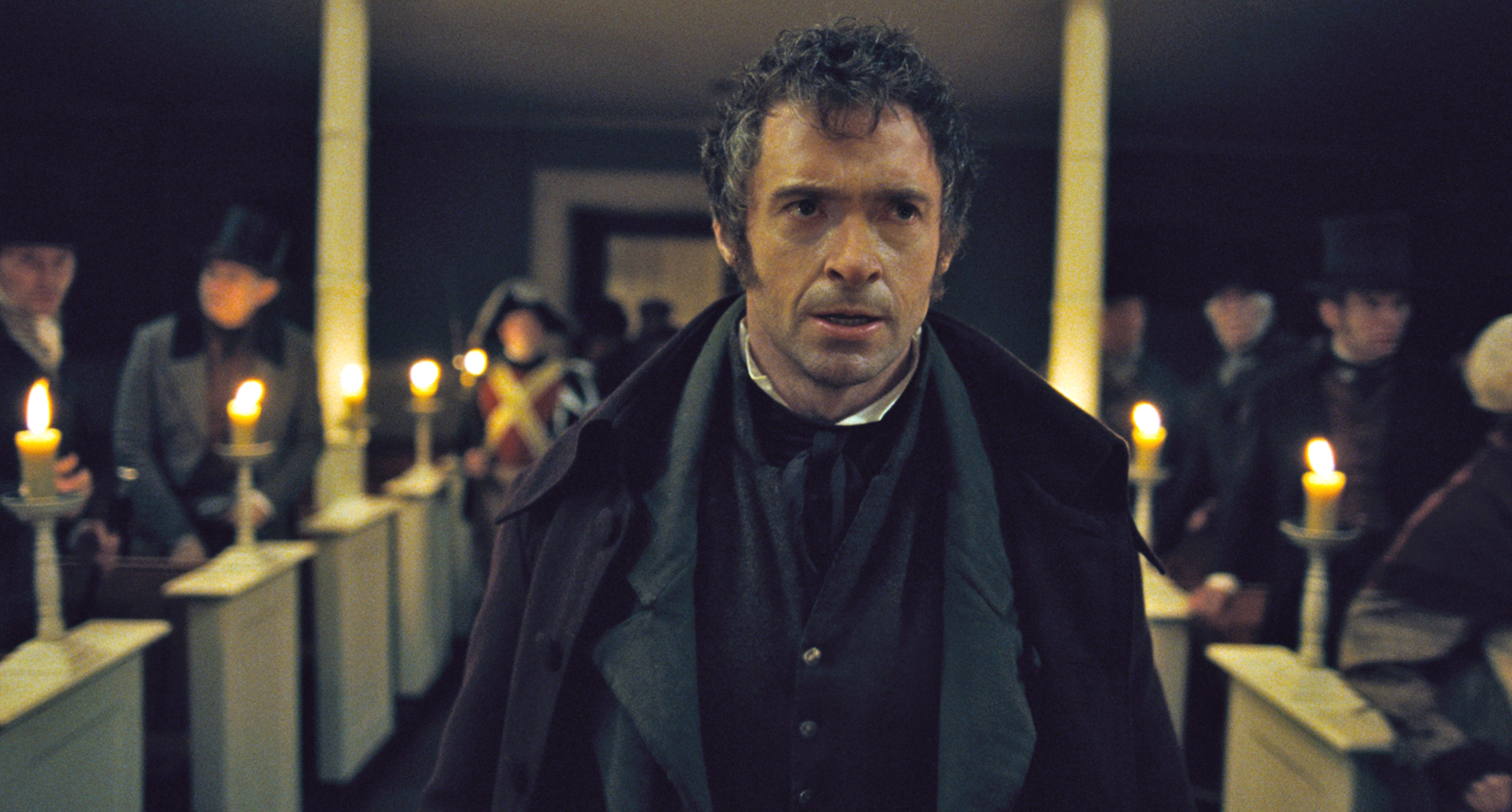 Hugh Jackman stands in a Victorian-era courtroom scene from a movie, wearing a period suit with a somber expression, surrounded by people holding candles