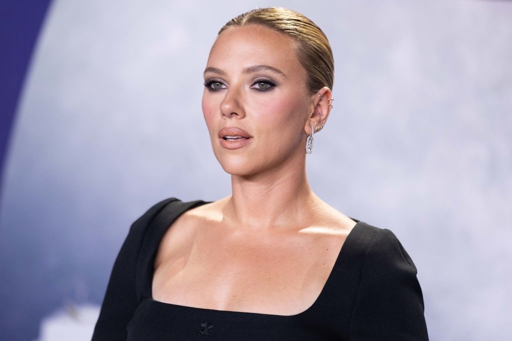 Scarlett Johansson on a red carpet, wearing a sleek, low-cut dress with long sleeves. She has her hair pulled back and is wearing drop earrings