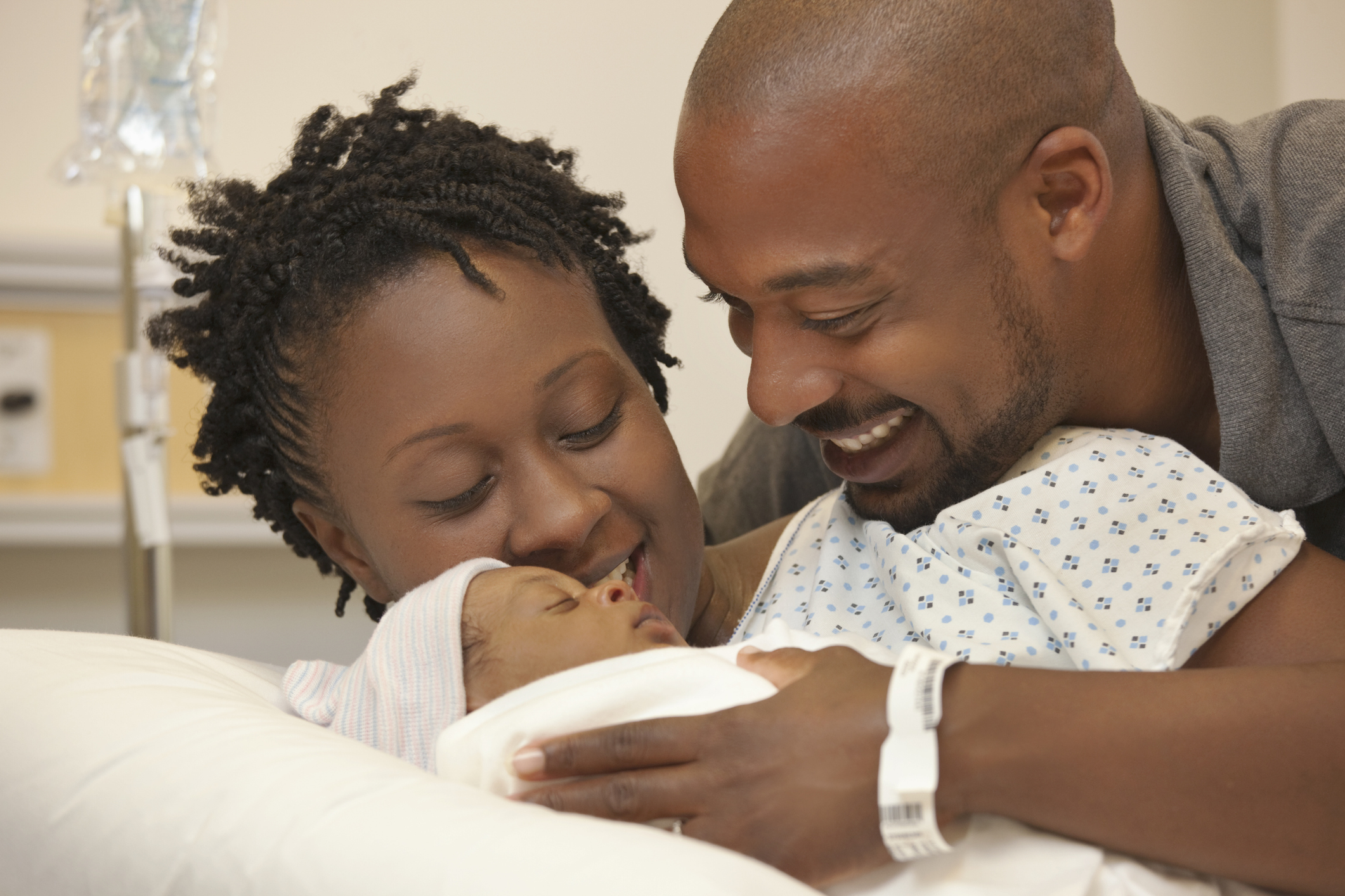 A loving couple smiles and admires their newborn baby in a hospital room. The father is wearing a patient wristband, while the mother gently cradles their child