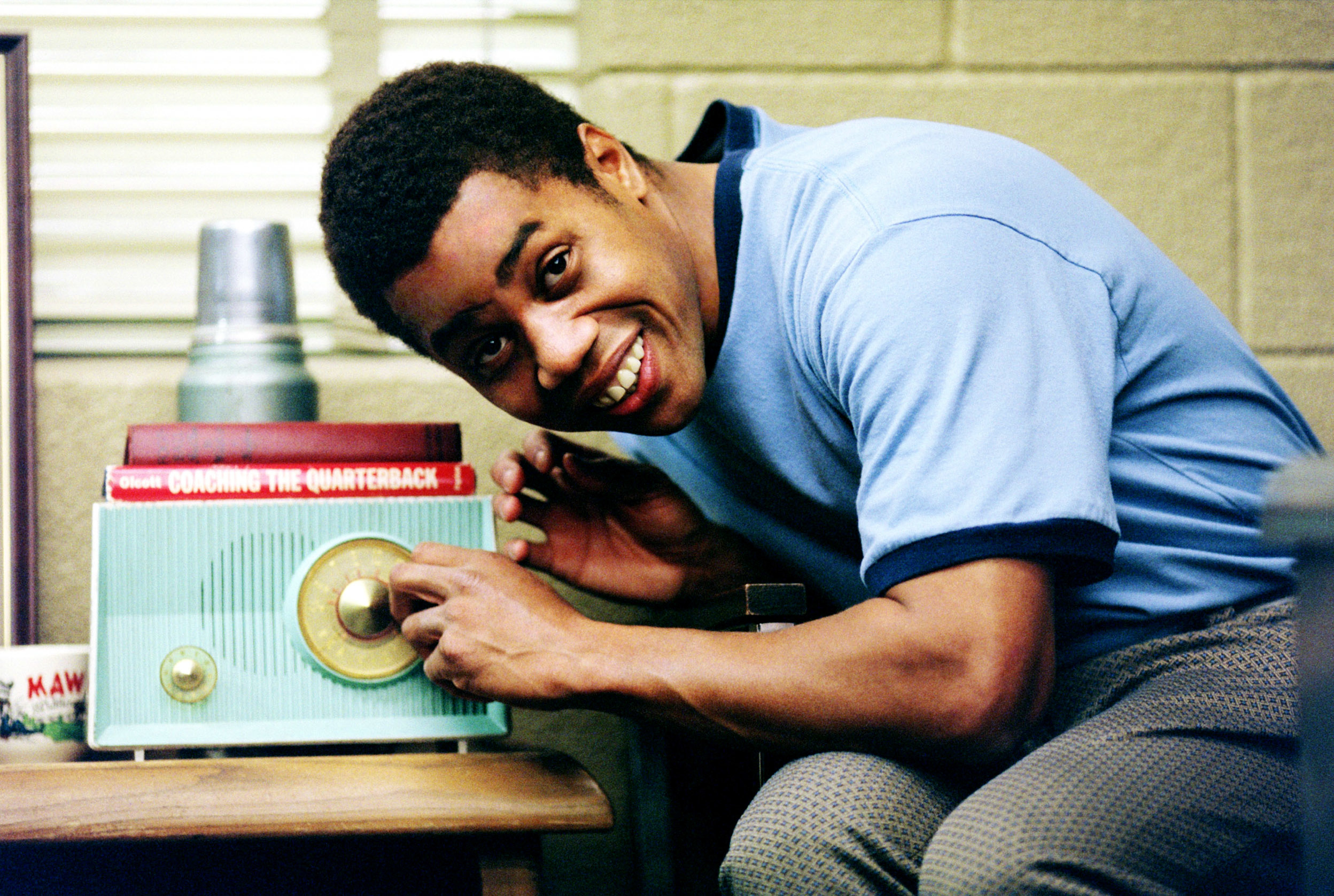 Cuba Gooding Jr., portrayed as Radio, smiles while tuning an old-fashioned radio with a stack of books in the background