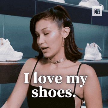Bella Hadid in a black spaghetti strap top, holding a pair of white sneakers in a shoe store, with text overlay saying "I love my shoes."