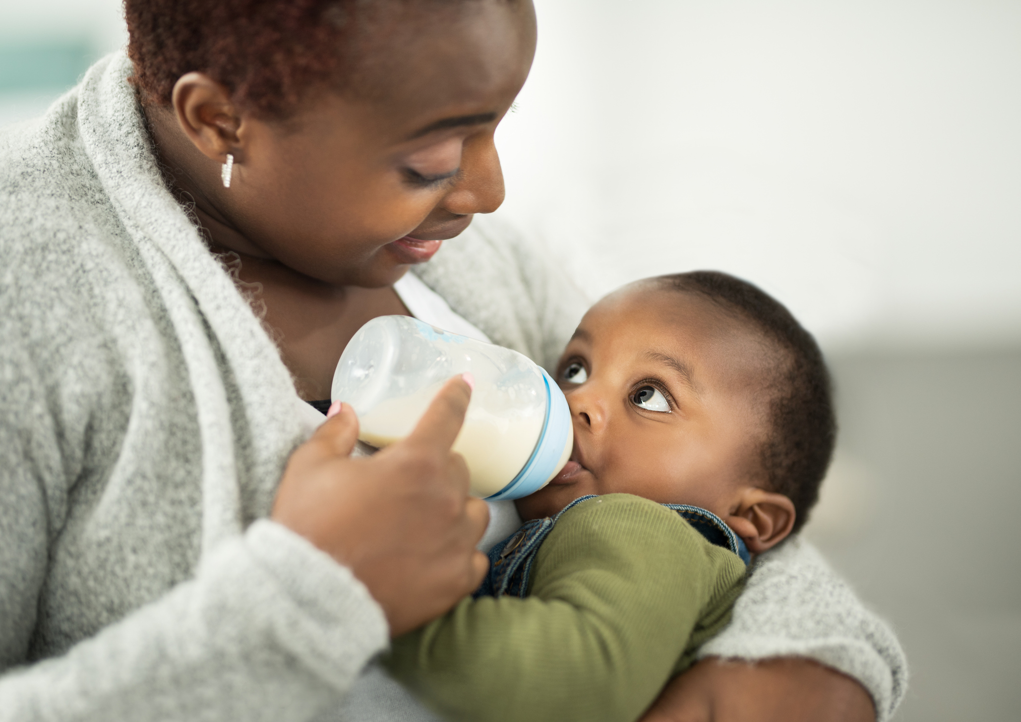 An adult woman feeds a baby with a bottle while holding them in her arms. Both are gazing at each other lovingly