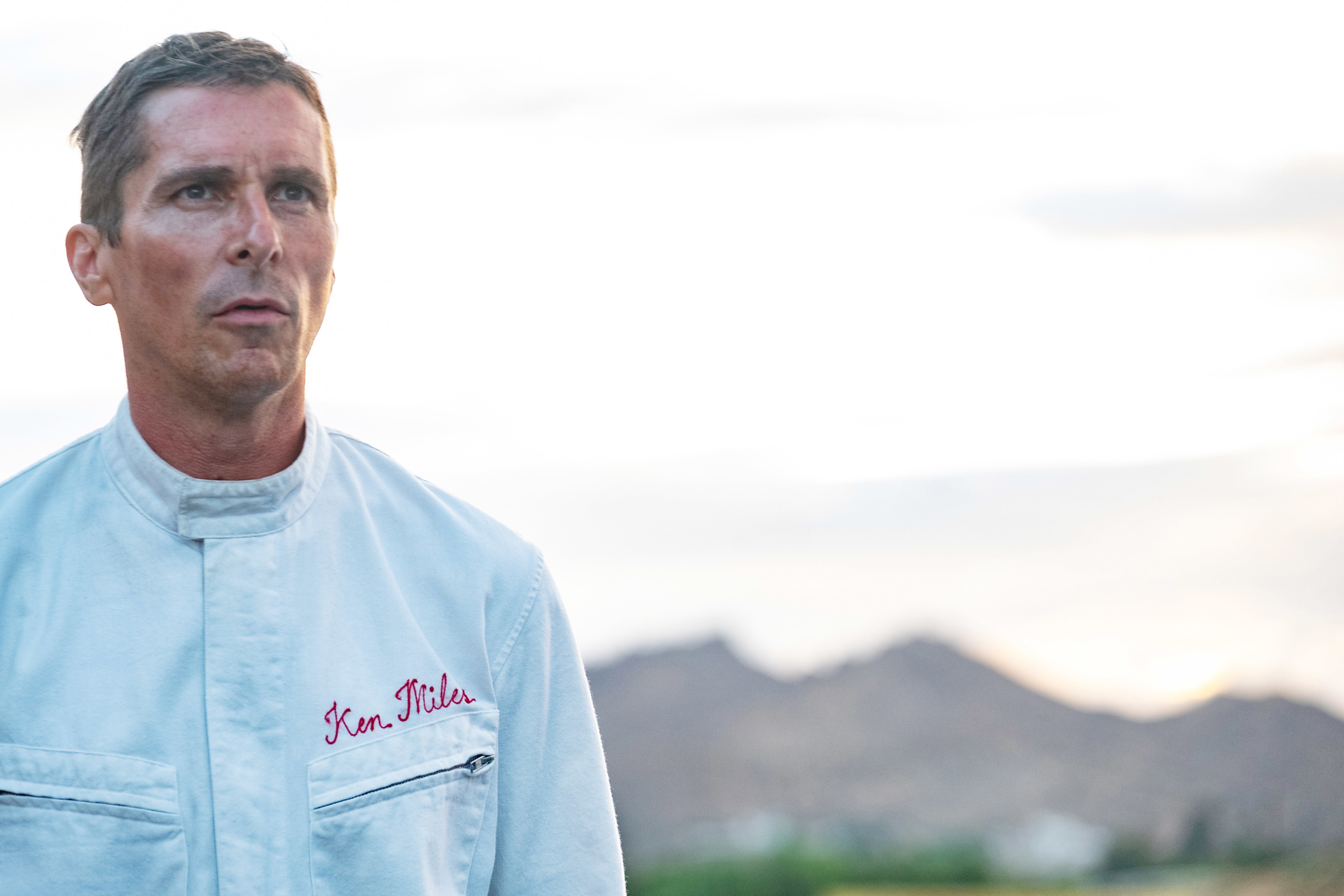 Christian Bale stands outdoors in a jacket embroidered with "Ken Miles," with blurred mountains in the background