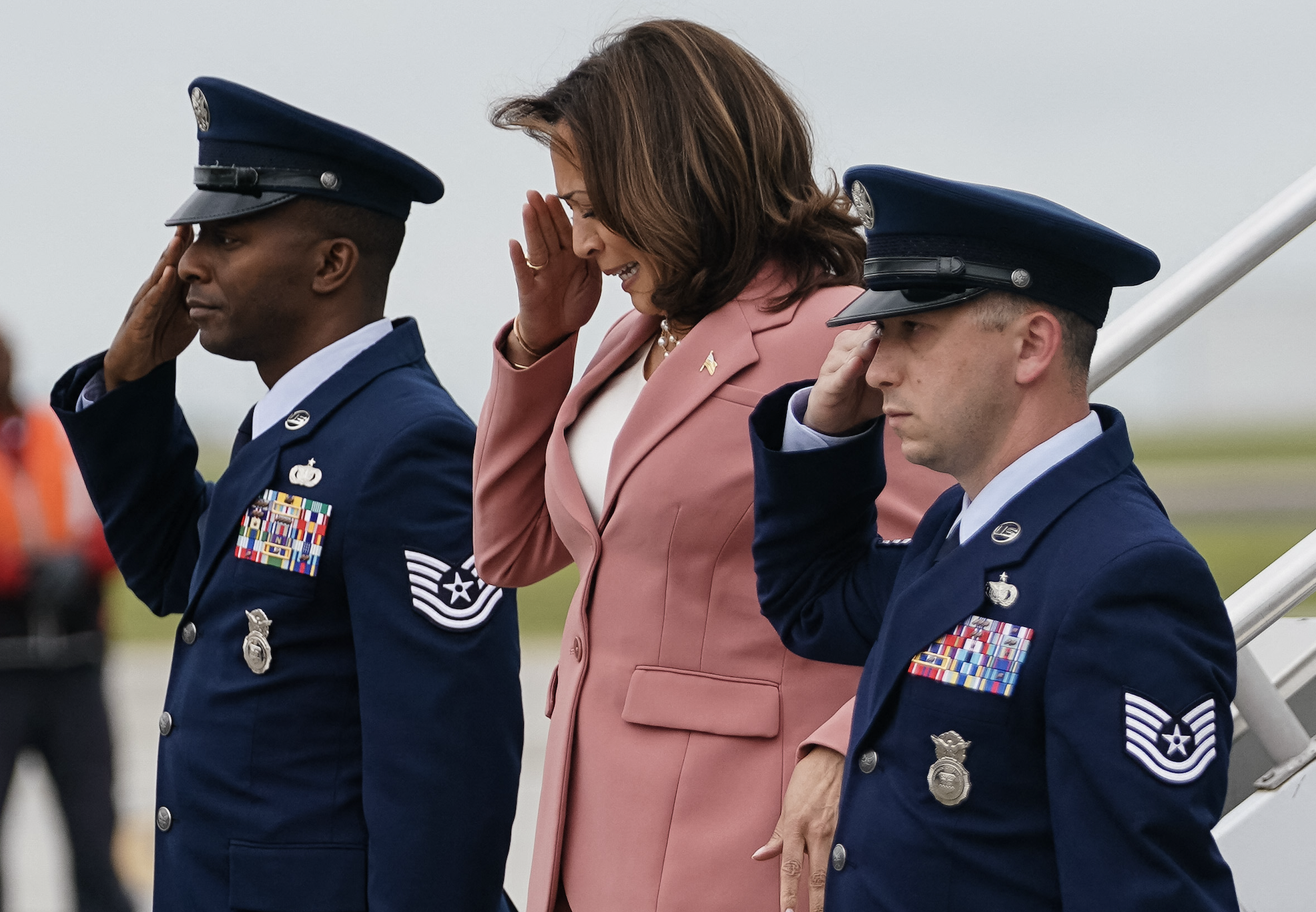 Kamala Harris salutes with two Air Force personnel in formal uniforms as they stand near an aircraft staircase