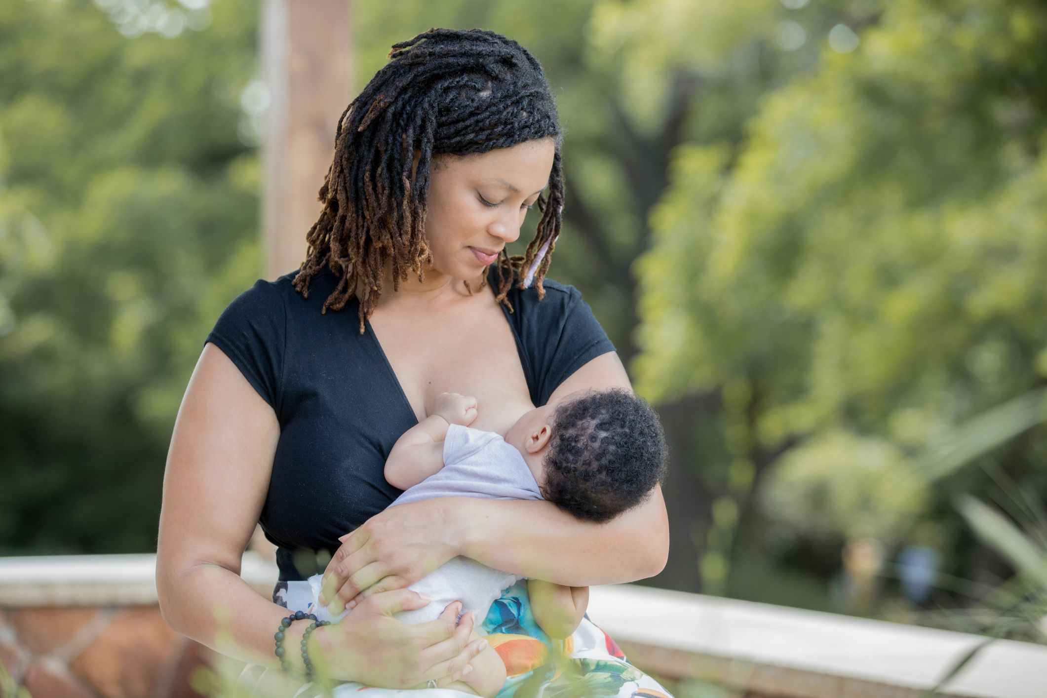 A woman with dreadlocks wearing a black top is breastfeeding a baby outdoors