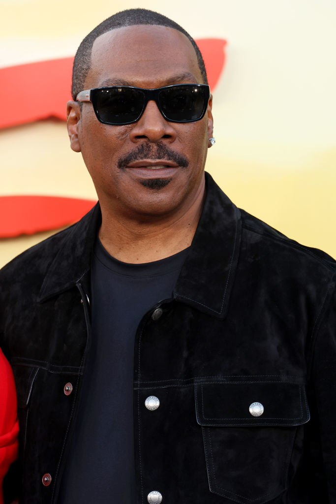 Eddie Murphy wears black sunglasses, a black shirt, and a black jacket at a public event