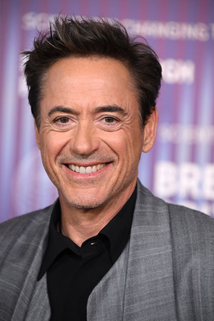 Robert Downey Jr. smiles for a photo on the red carpet, wearing a grey blazer over a black shirt