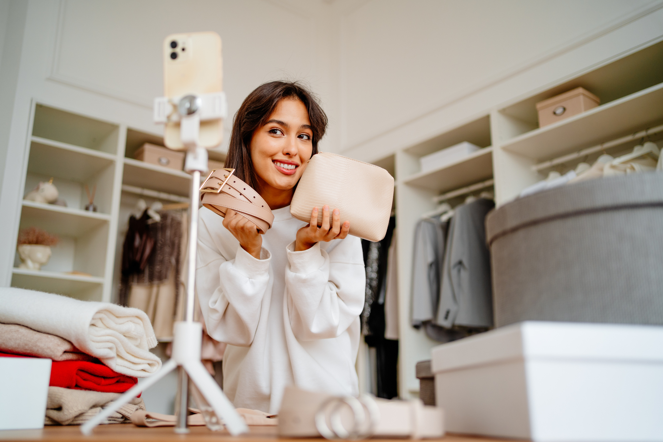 Woman in a closet holds a purse and a belt while smiling at a smartphone on a tripod
