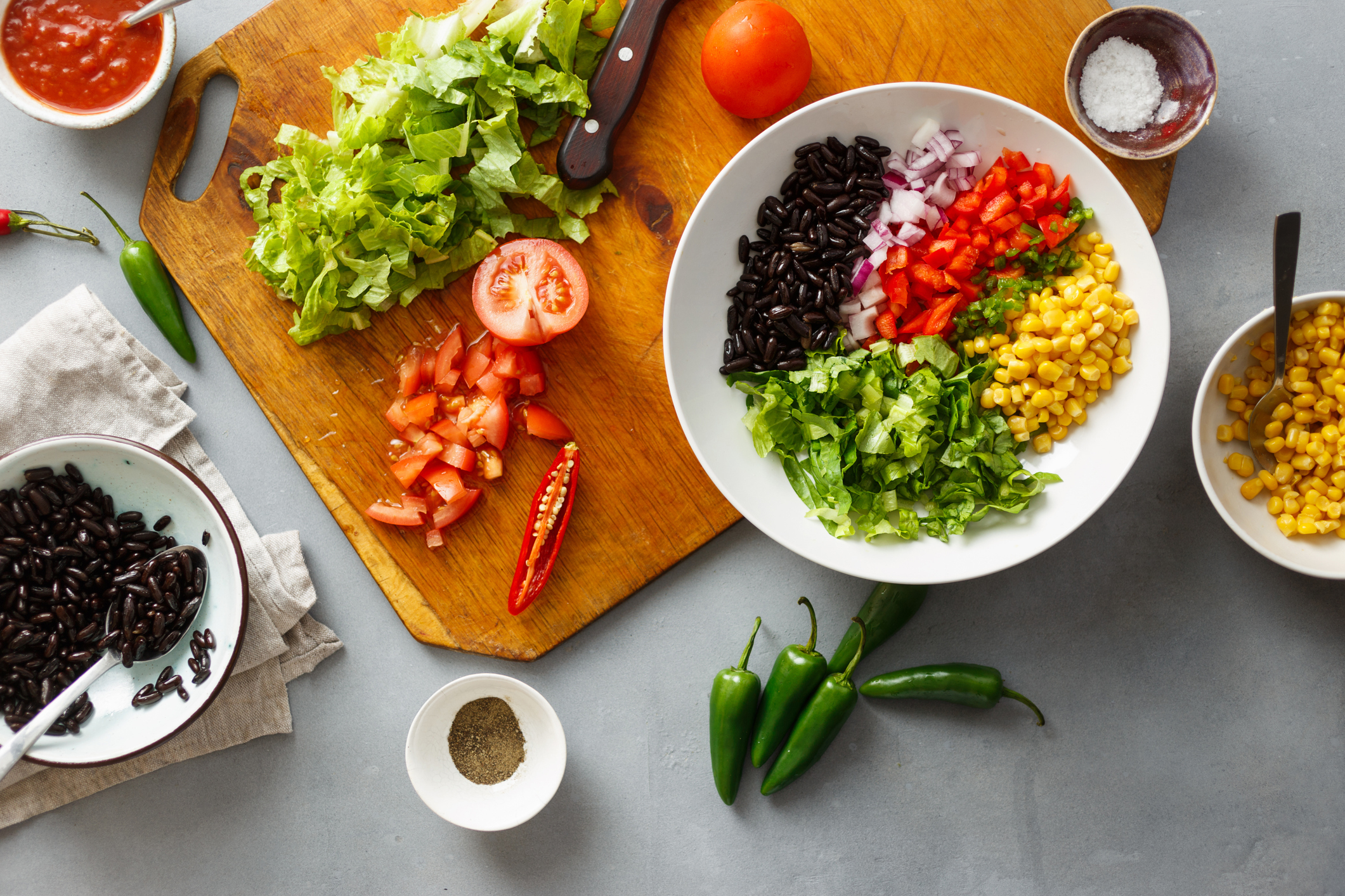 A cutting board with chopped vegetables next to a bowl containing black beans, diced onions, red peppers, corn, and lettuce. Various ingredients surround the board