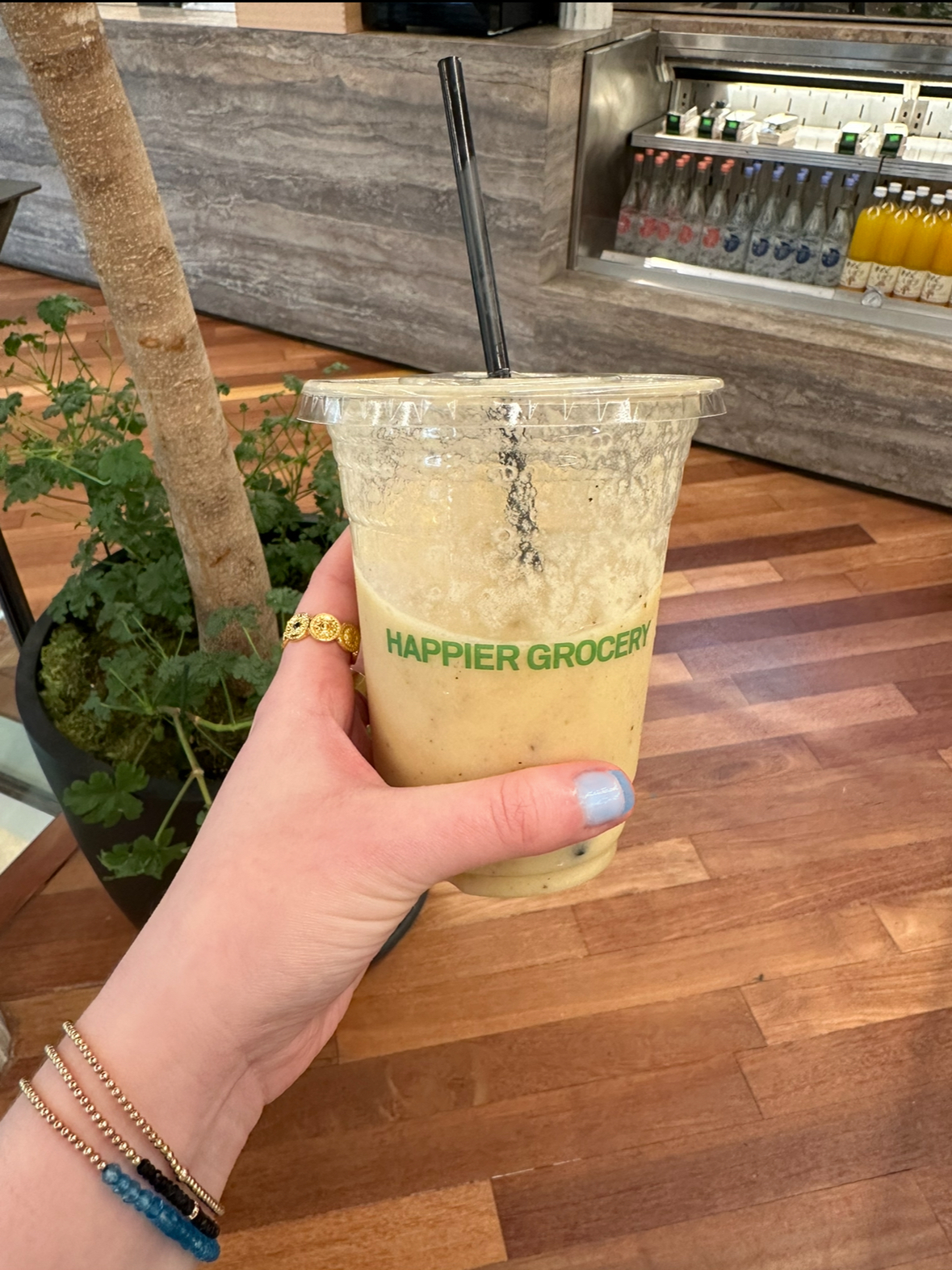 A hand with bracelets holds a smoothie in a plastic cup with "Happier Grocery" printed on it inside a store