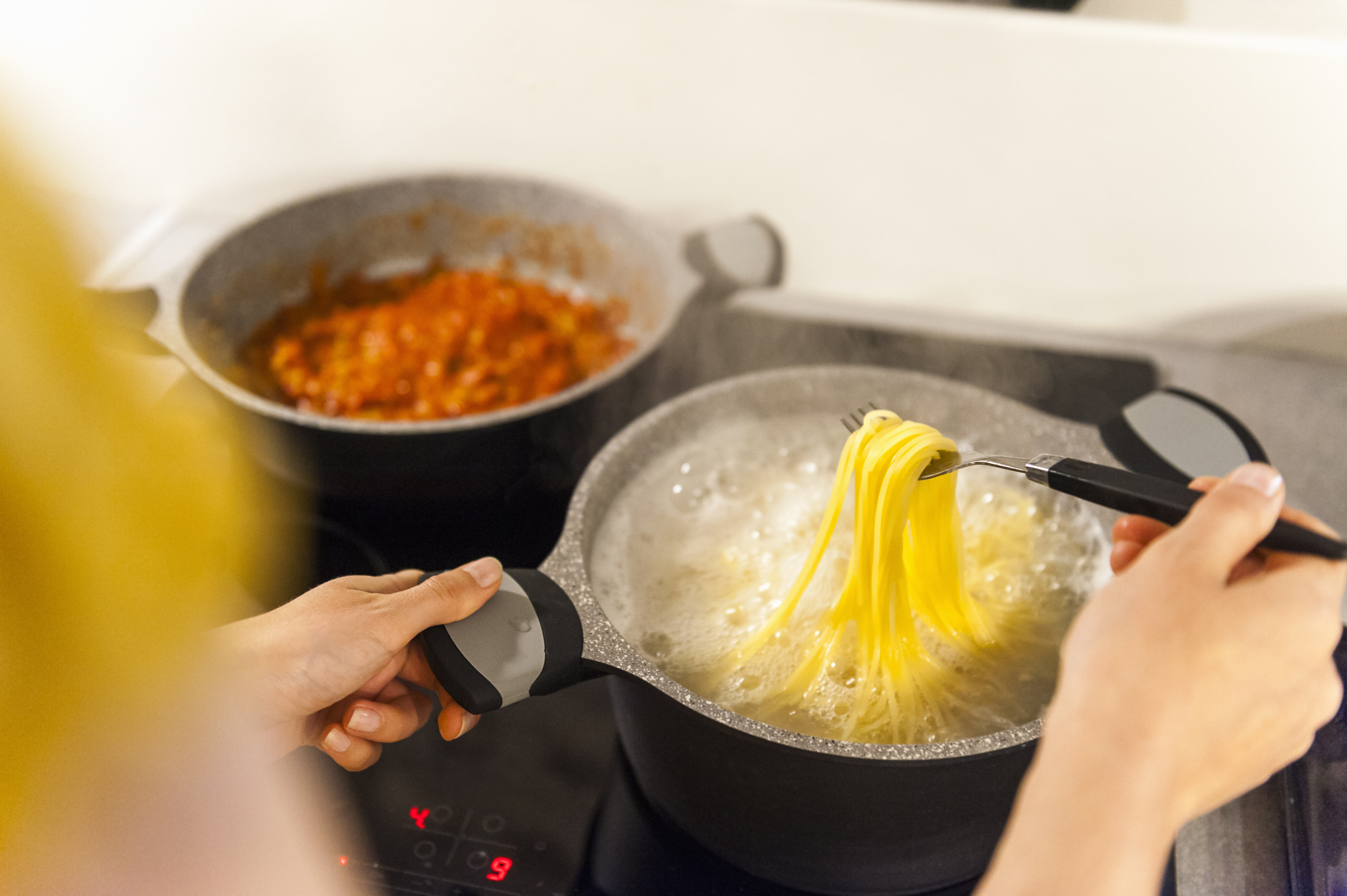 A person is cooking pasta in a pot while another pot with a red sauce simmers in the background