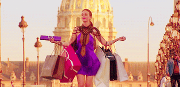 Blair Waldorf in a stylish dress walks outdoors, carrying multiple shopping bags in both hands. She is smiling and appears joyful