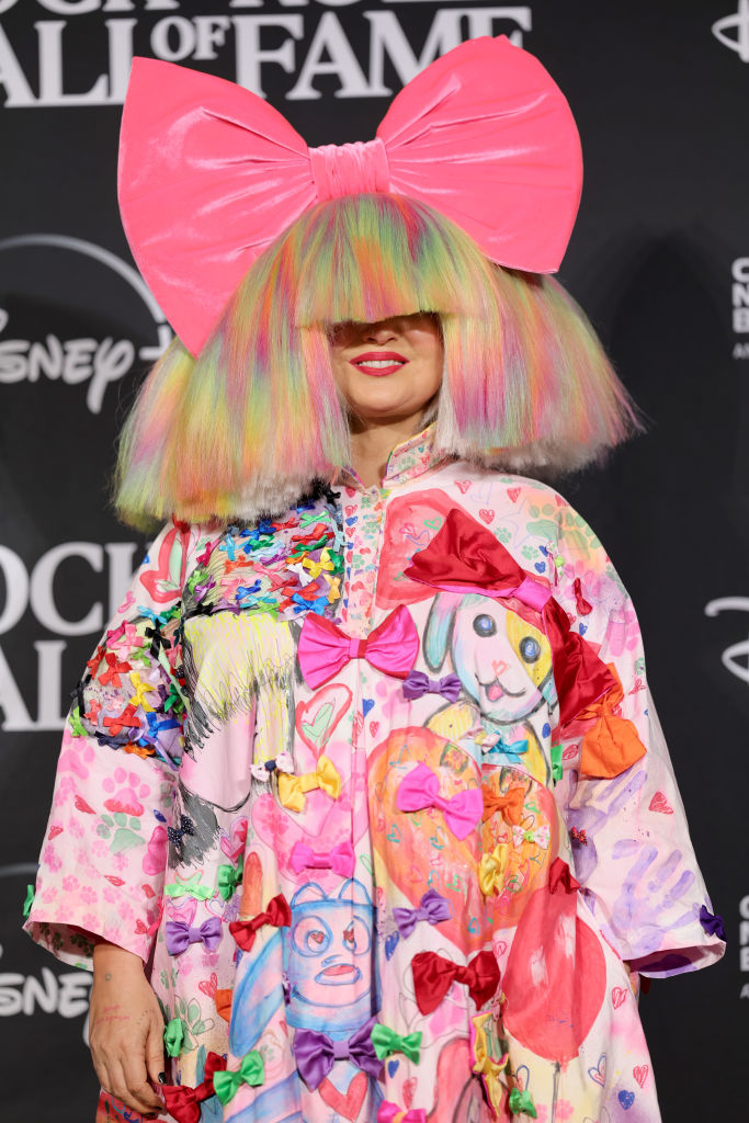 Sia on the red carpet, wearing a colorful outfit with child-like drawings and a large pink bow in her hair, covering her eyes