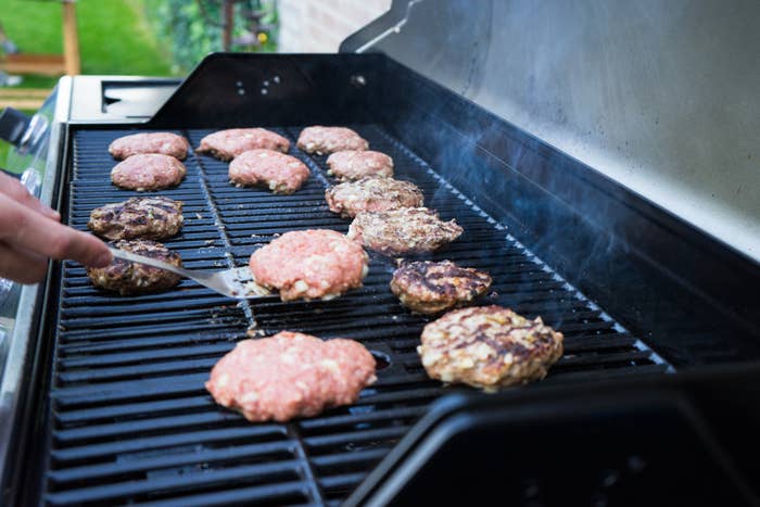 Several patties cooking on a barbecue grill, with a hand flipping one patty using a spatula. Steam rises from the grill