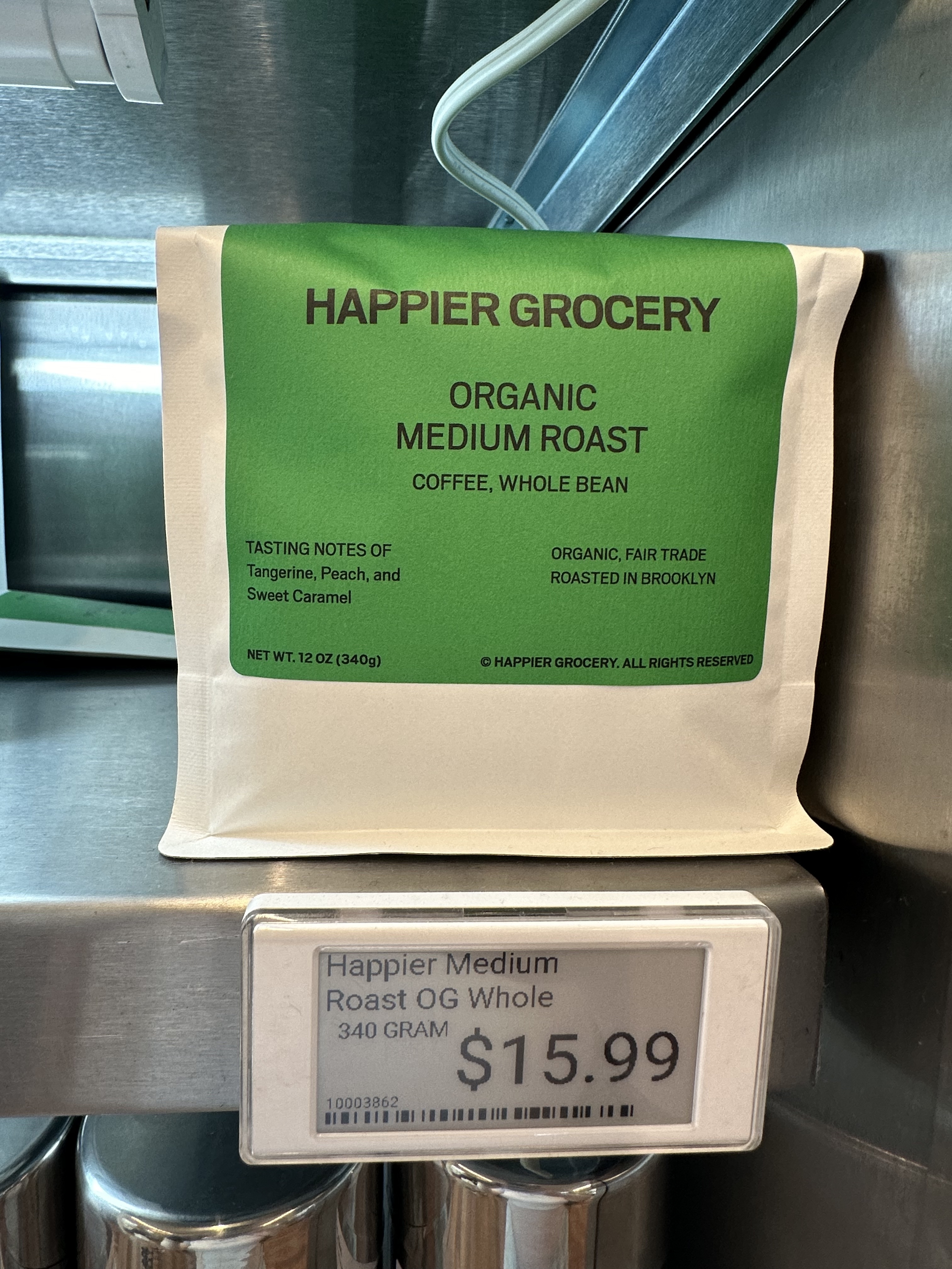 Happier Grocery Organic Medium Roast whole bean coffee on a shelf, priced at $15.99 for 340 grams. Notes of tangerine, peach, and sweet caramel.