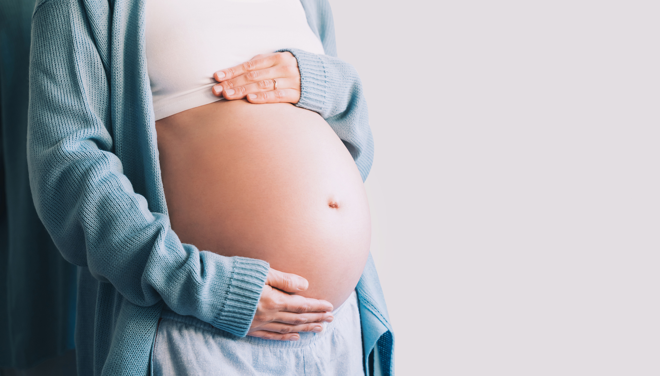 Pregnant person wearing a white top and cardigan, cradling their belly with both hands