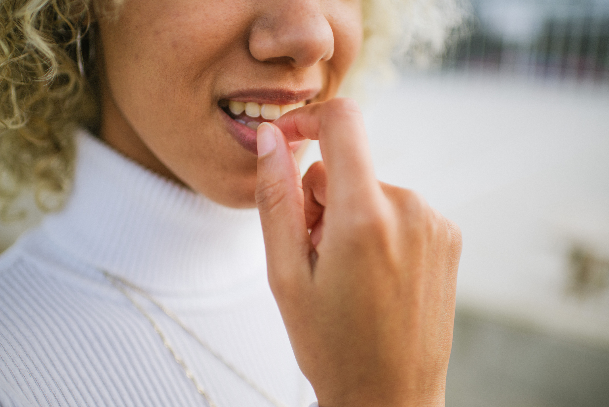 Close-up of a person with light curly hair, touching their lips with their finger. Wearing a white turtleneck sweater and a necklace