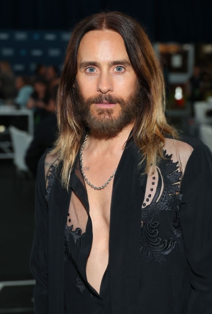 Jared Leto is shown wearing a partially unbuttoned shirt with intricate designs and a chain necklace, standing in what appears to be a formal event setting