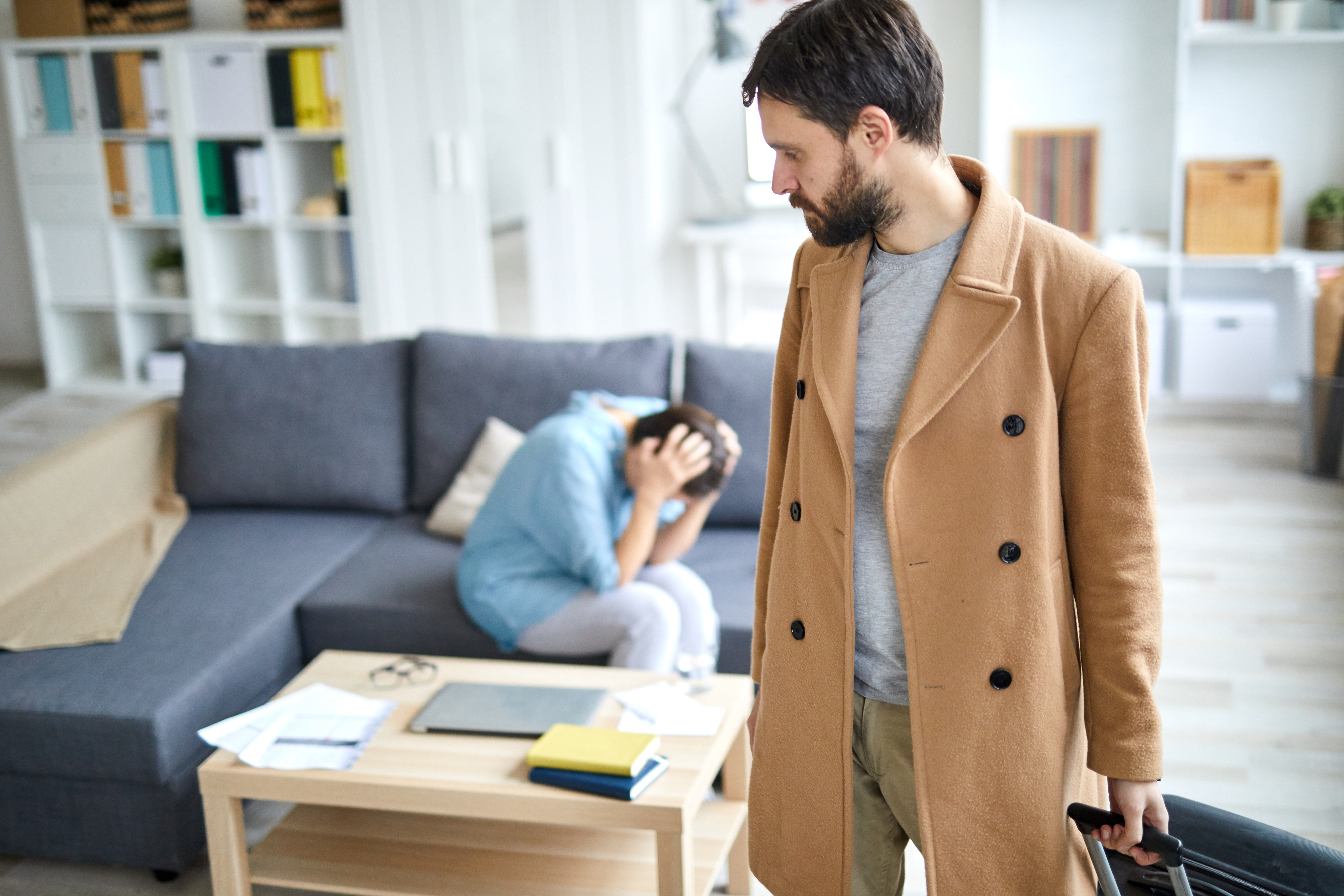 A man in a coat holding a briefcase looks back at a distressed person on the couch, who has their head in their hands amidst scattered papers