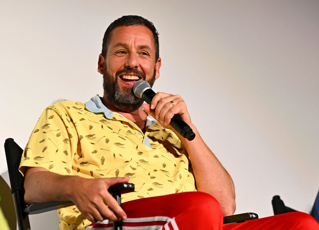 Adam Sandler, wearing a polo shirt and red pants, sits in a chair holding a microphone and smiling